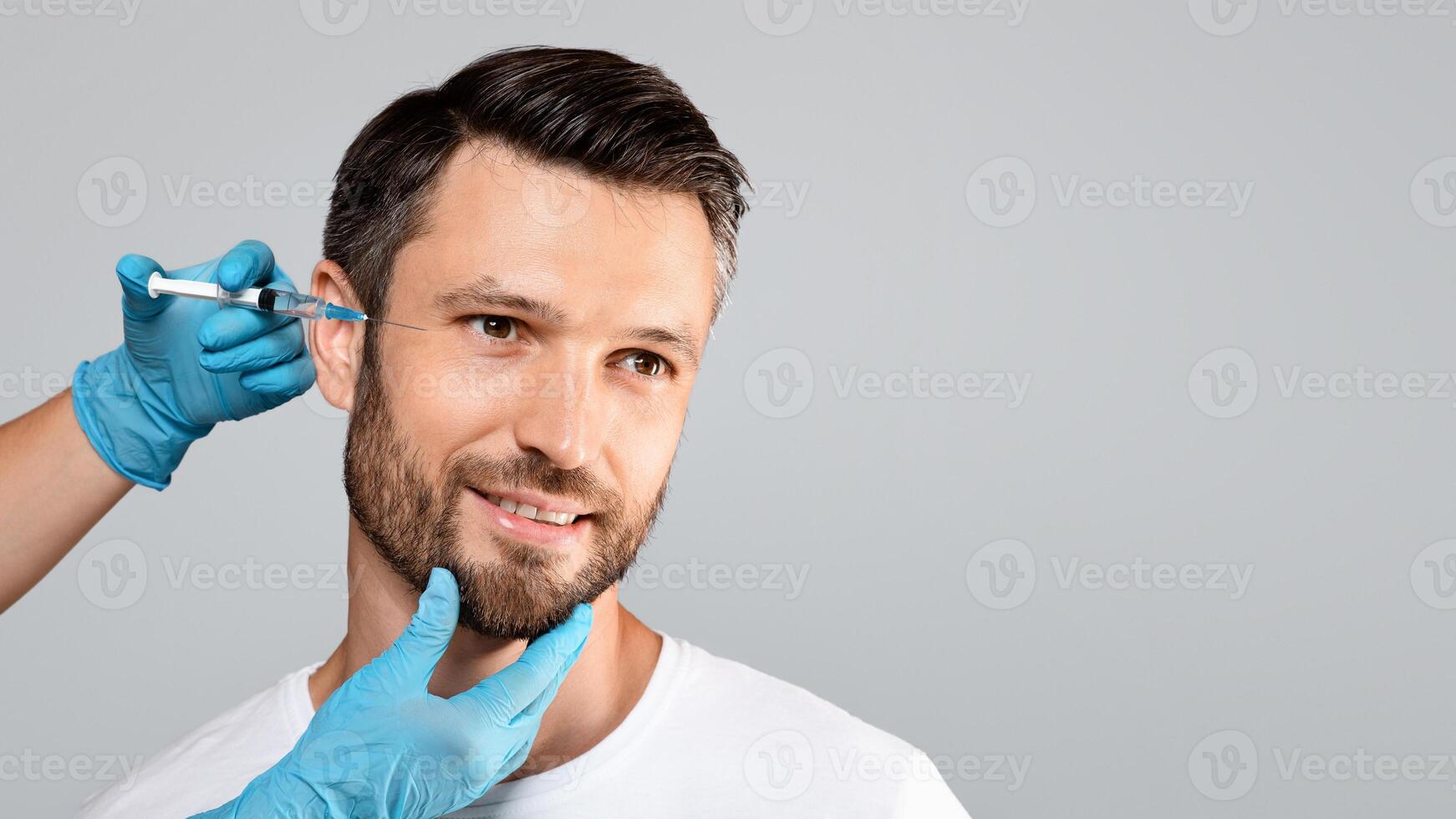 Handsome bearded man getting under eye injections in beauty clinic, looking at copy space, grey studio background. Middle-aged smiling man attending beautician, having face filler photo