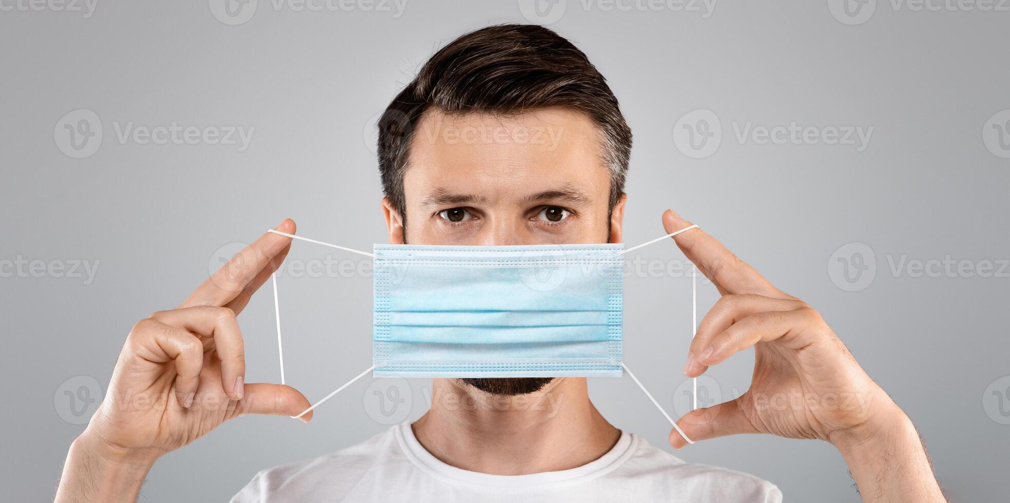 Bearded man holding medical protective mask on grey studio background. Man in white t-shirt with blue protective medical mask, healthcare concept, stay safe and healthy during pandemic photo