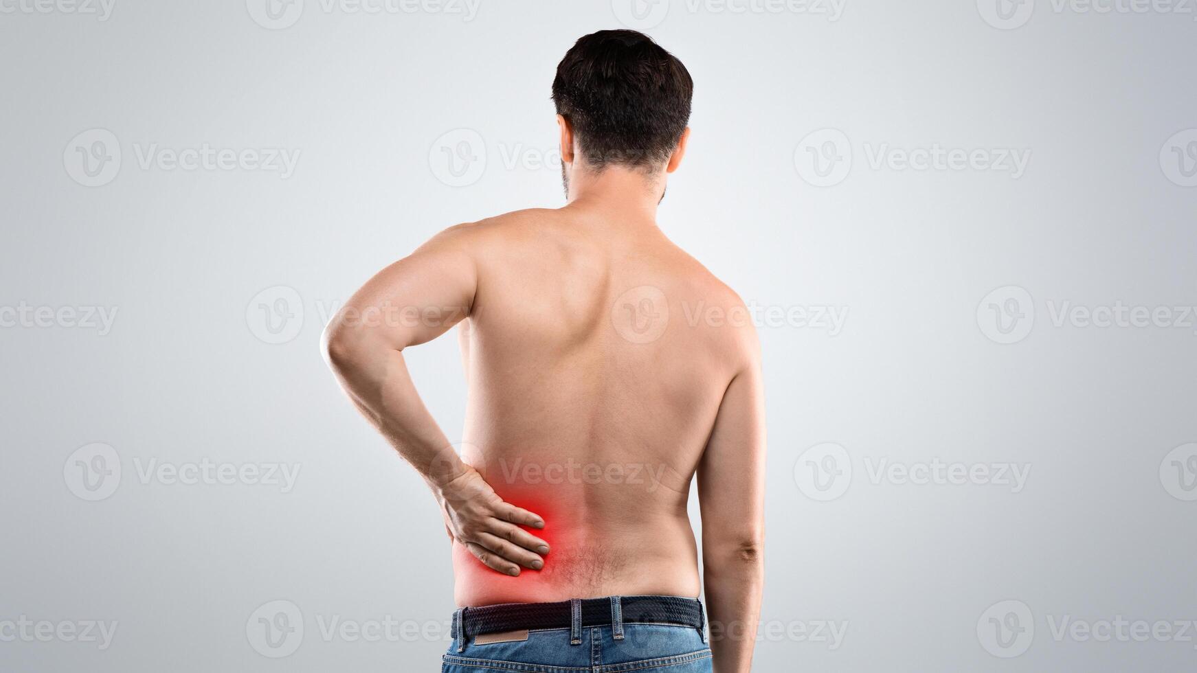 Back view of unrecognizable topless man touching his lower back zone highlighted with red, suffering from chronic or acute kidney disease, grey studio background, empty space. Healthcare concept photo