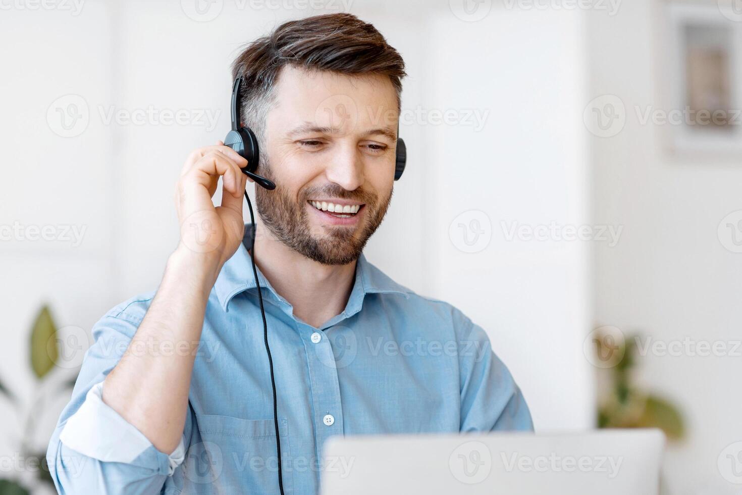 Hotline Operator. Handsome Young Man Customer Support Manager In Headset Working With Laptop Computer In Office, Consulting Clients Online, Managing Issues, Sitting At Desk, Free Space photo