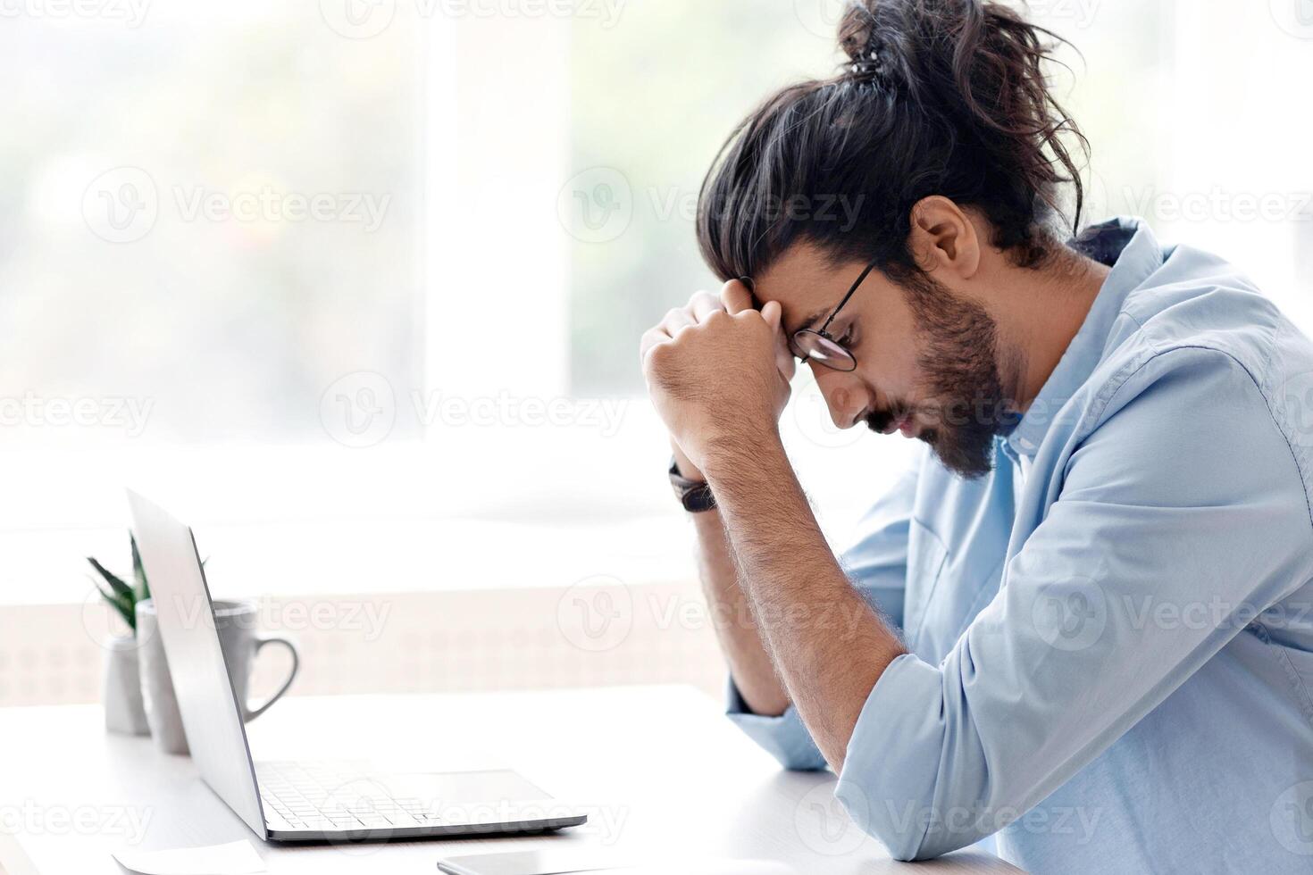 Upset Eastern Freelancer Guy Suffering Lack Of Inspiration While Working On Online Project With Laptop At Home, Sitting At Desk Near Window And Touching Head In Despair, Side View With Copy Space photo