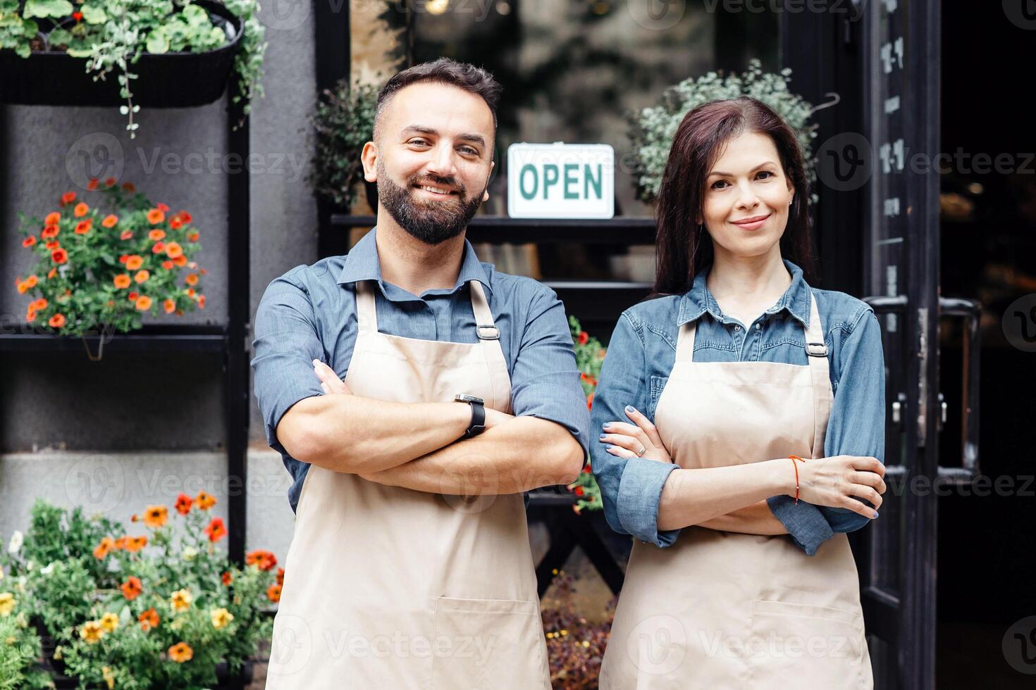 Opening after covid-19 virus, small business and floral design. Cheerful millennial guy and lady in aprons with crossed arms standing near store with different plants in pots and sign is open, outdoor photo