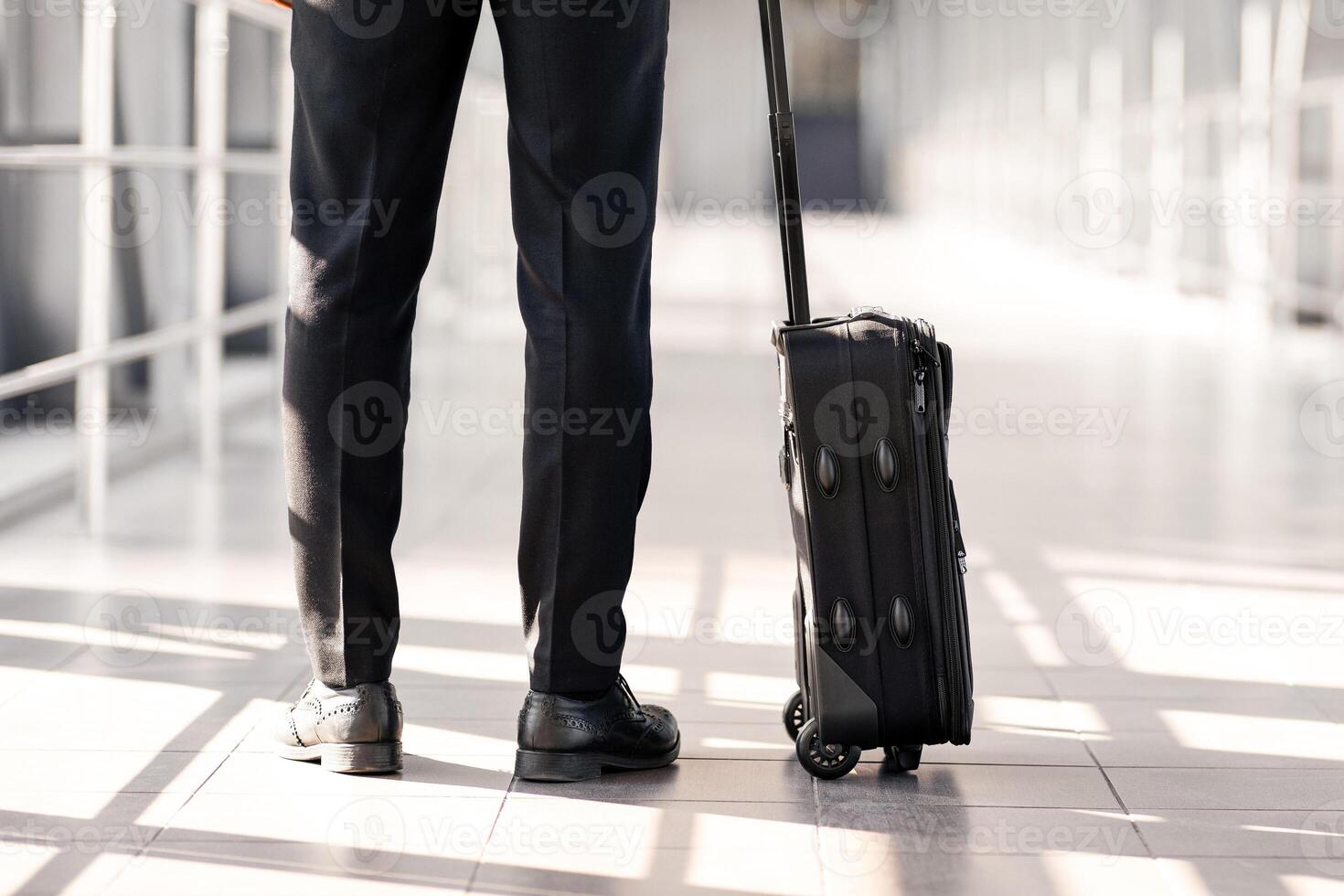 Cropped of businessman in black suit with suitcase standing in airport, back view, panorama with copy space. Unrecognizable man having business trip, going for departure or arrived for meeting photo