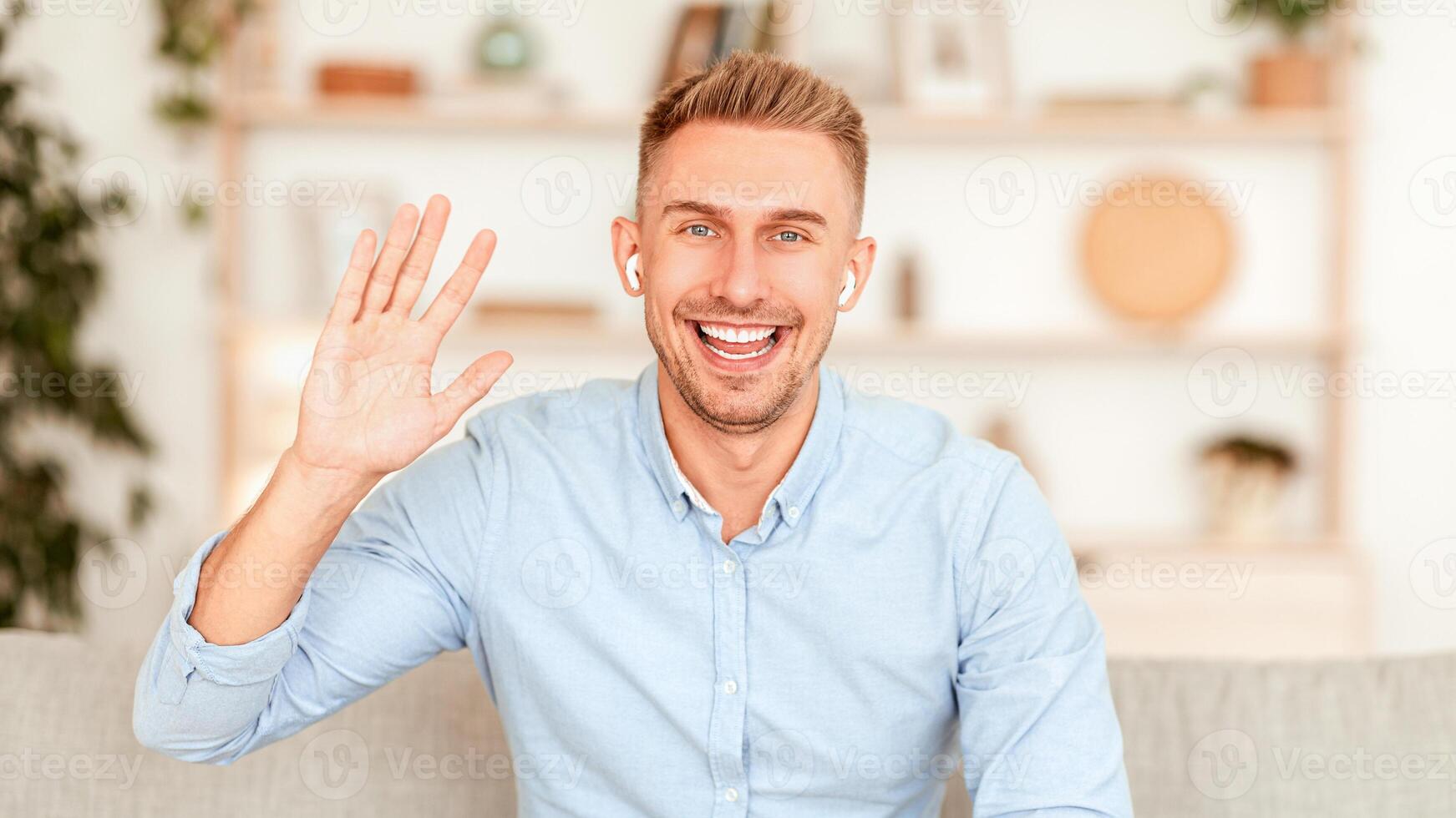 Hi. Positive Millennial Guy Waving Hello With Hand Smiling To Camera Sitting On Couch At Home, Wearing Earbuds photo