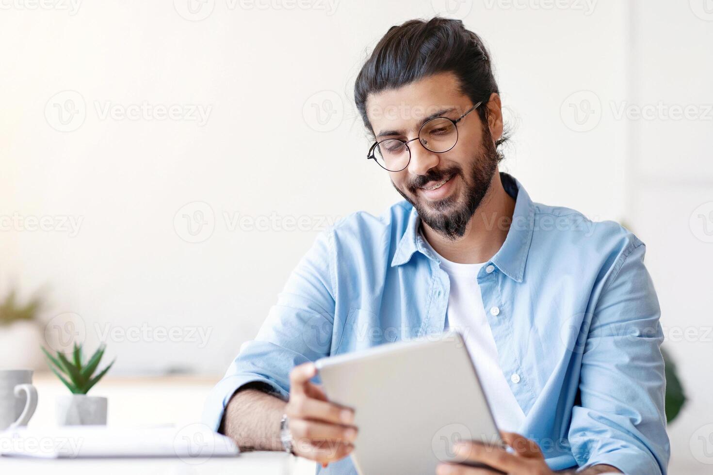 Arab Freelancer Guy Using Digital Tablet At Home, Having Break In Work, Sitting At Desk, Browsing Internet Or Social Media, Checking New App, Smiling Eastern Man Messaging Or Shopping Online photo