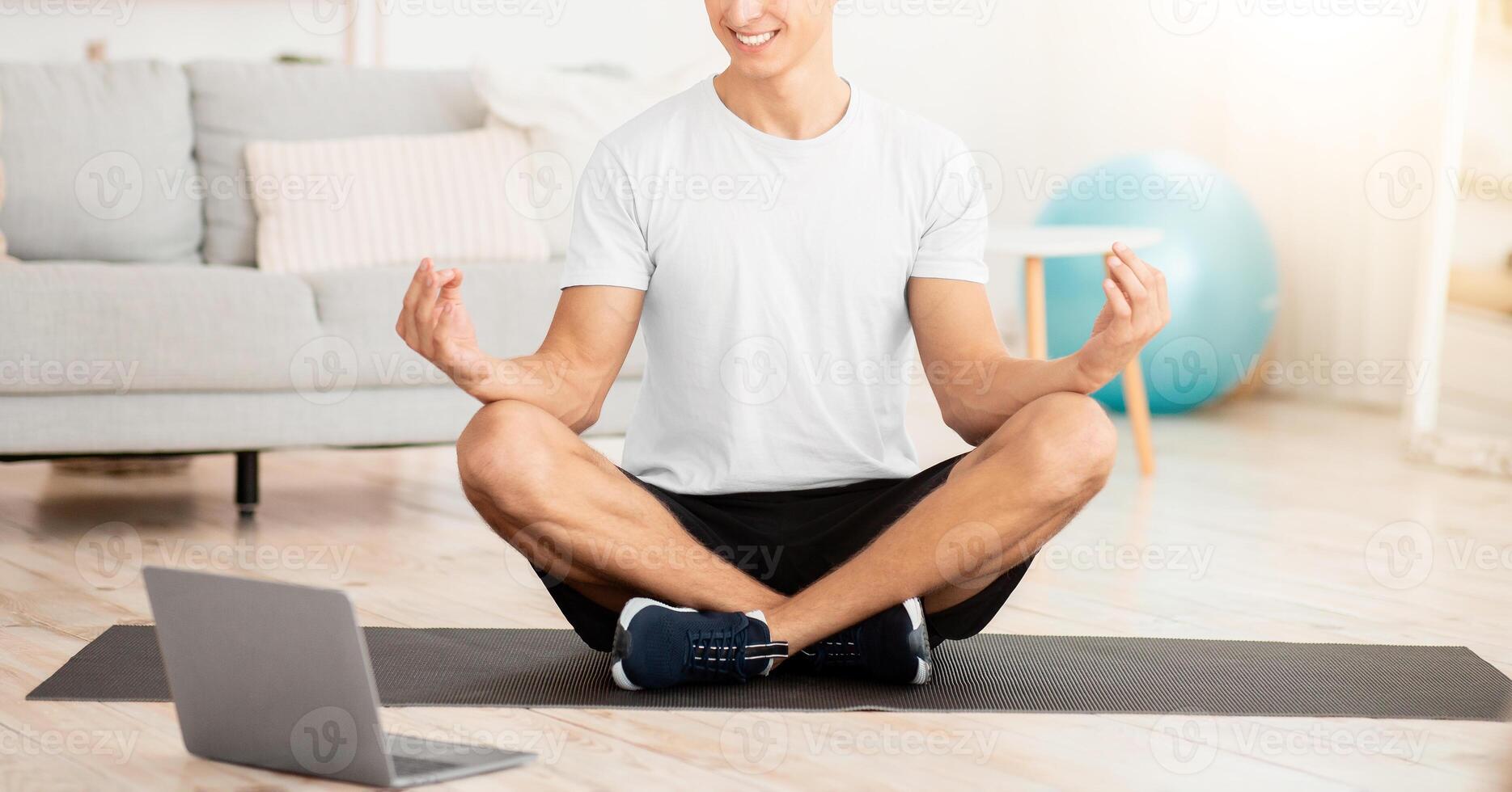 Online yoga course. Laughing young man in sportswear sitting in lotus position and trying to meditate on mat in interior of living room, panorama, free space photo