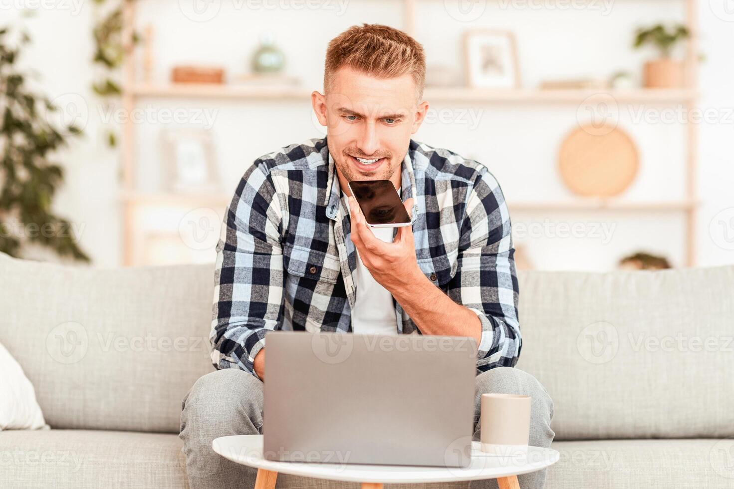 Voice Search. Portrait of guy talking on smartphone or using virtual personal assistant app on his device sitting on the couch in living room, working on laptop. Man recording audio message or command photo