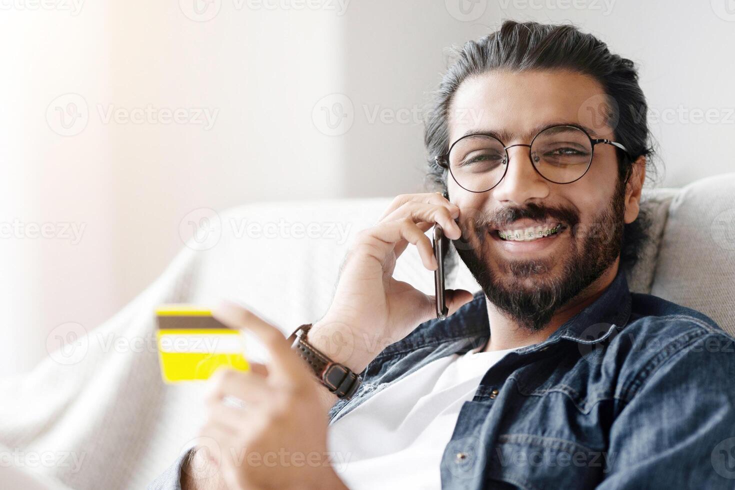 Handsome Young Arabic Man Holding Credit Card And Talking On Cellphone At Home, Enjoying Making Easy Online Payments, Speaking With Customer Support While Relaxing On Couch In Living Room, Free Space photo