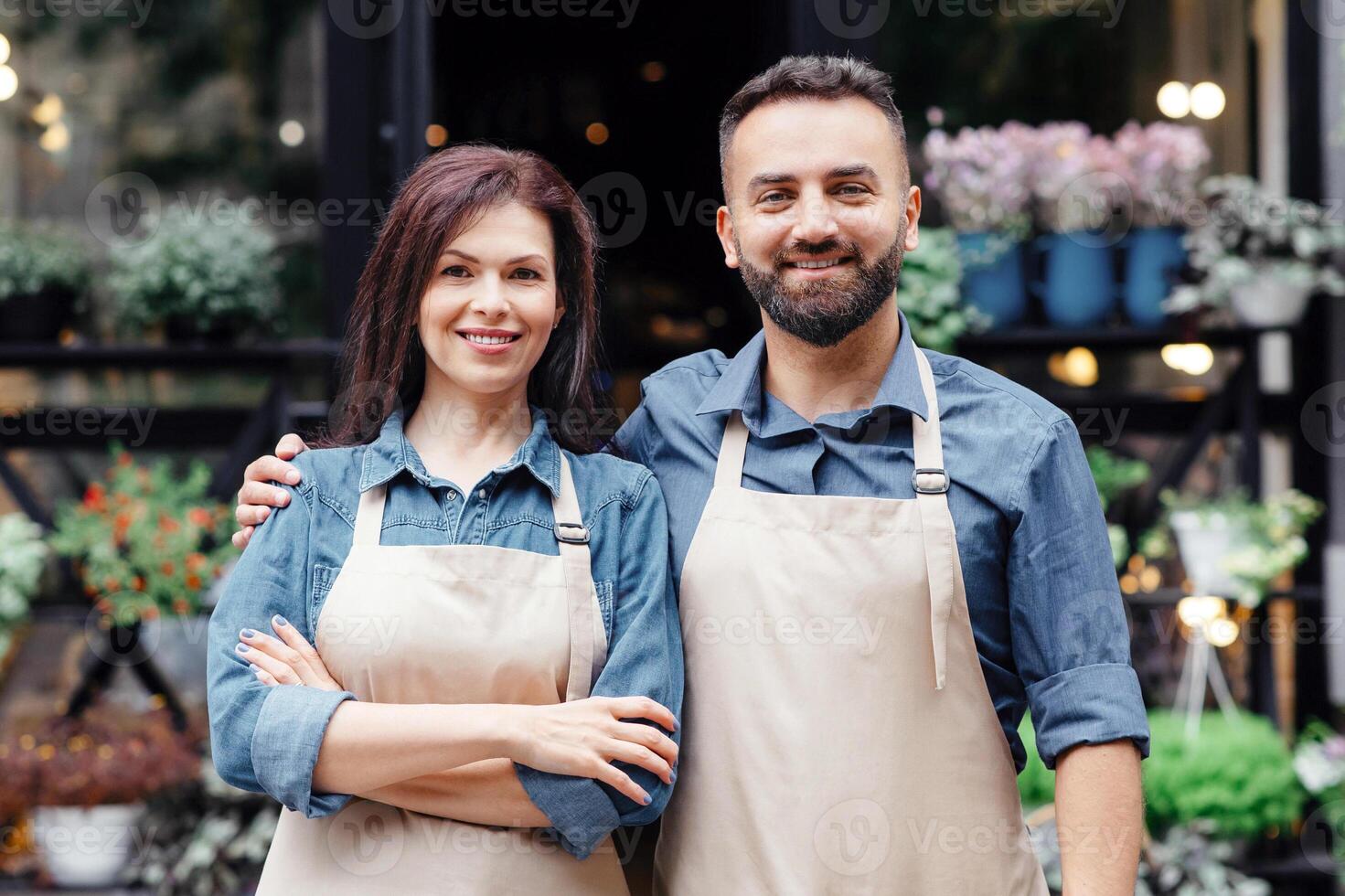 Owners of flower shop near showcase outdoor. Happy millennial bearded european guy and lady in aprons waiting clients in studio or eco cafe interior in rustic style with different plants, empty space photo