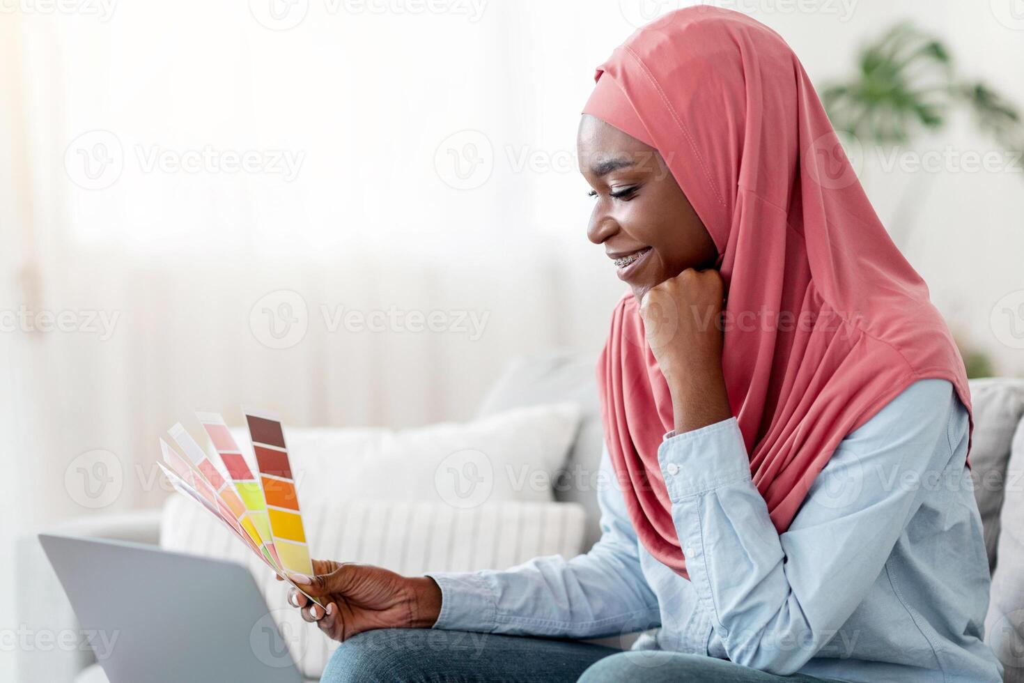 Smiling Black Muslim Lady Holding Color Swatch And Using Laptop At Home, Choosing Interior Design For New Apartment, Sitting On Couch In Living Room, Checking Papers With Colour Palette, Copy Space photo