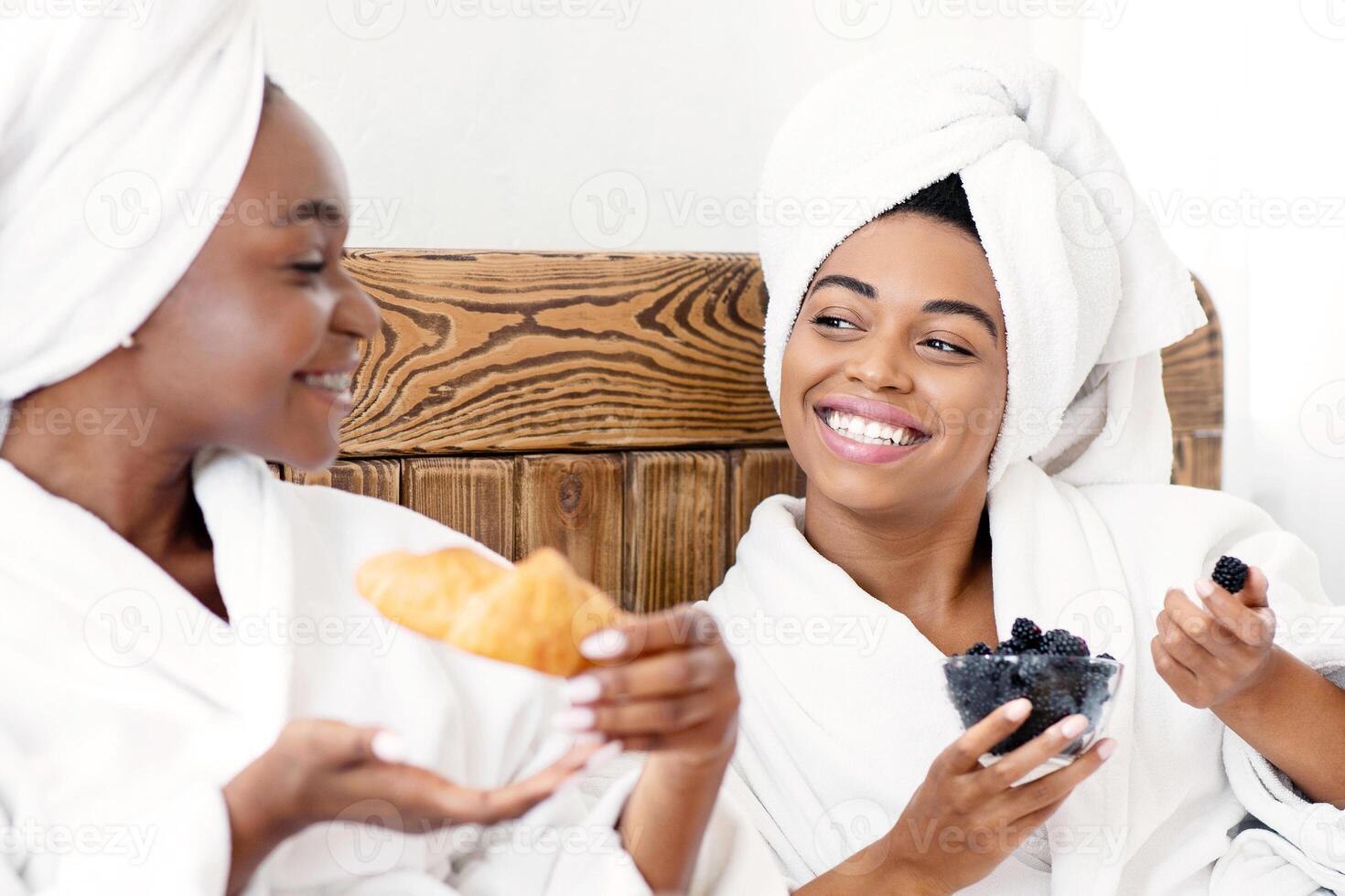 Closeup of beautiful african american ladies having healthy snack in bed, having relaxing spa day together at home. Two cheerful black women enjoying breakfast, talking and laughing, bedroom interior photo