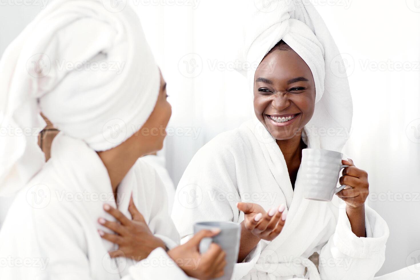 Two joyful african american girlfriends in bathrobes joking and drinking coffee. Cheerful black women with towels on their head having spa day at home, drinking tea and having funny conversation photo