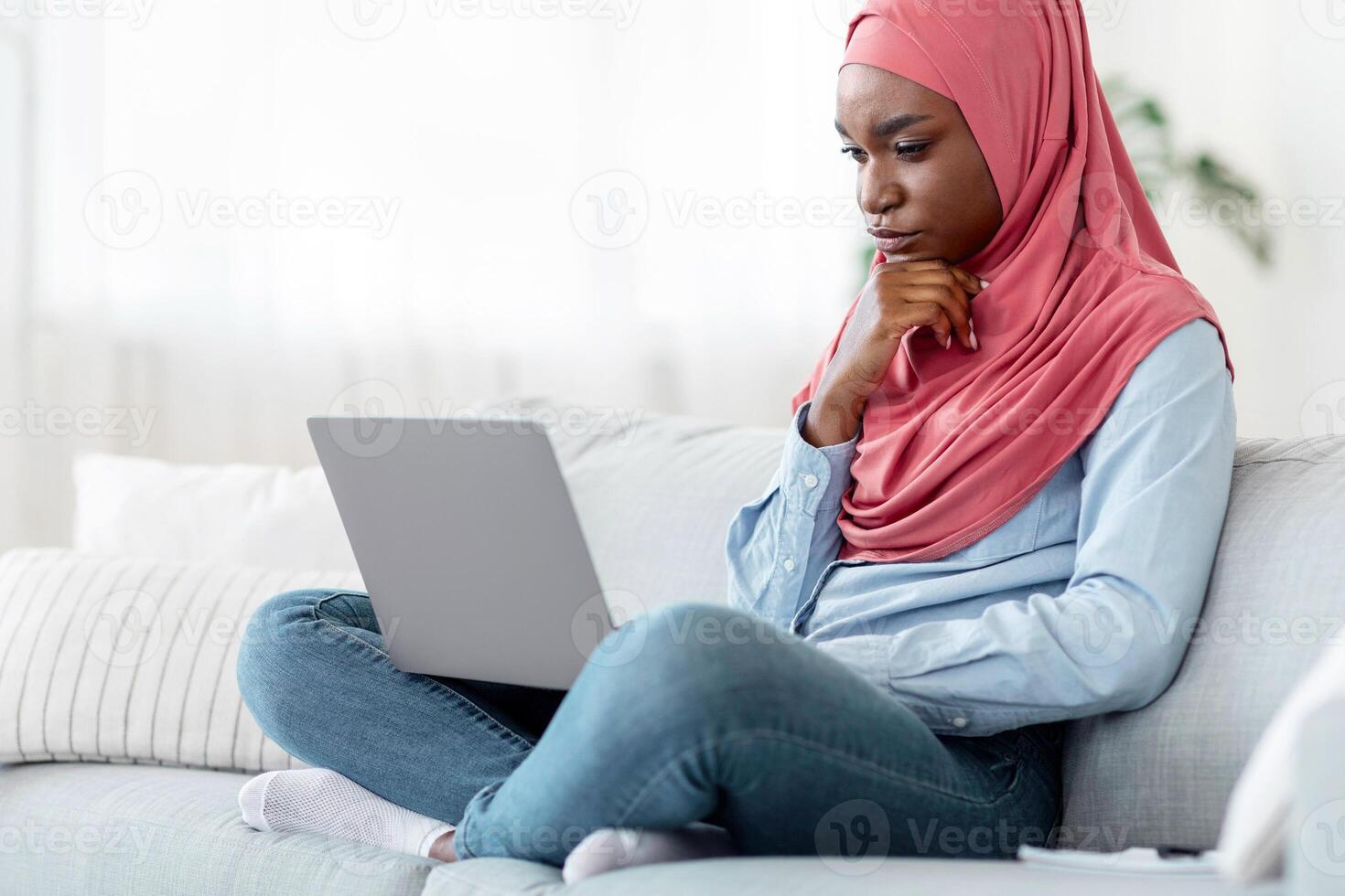 Pensive black muslim female freelancer with laptop at home brainstorming at new project, making research in internet, having remote job, sitting on couch in living room looking at computer screen photo