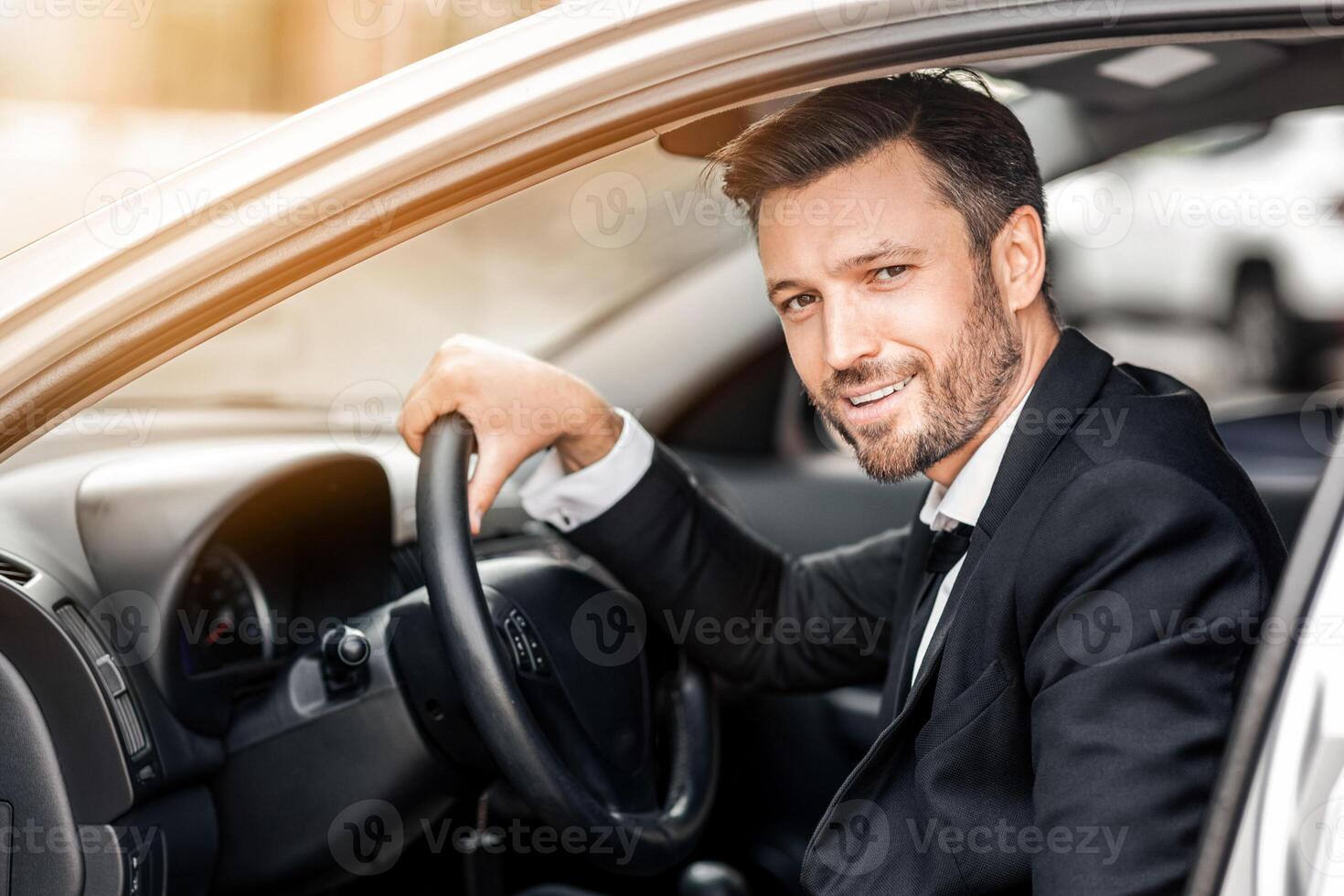 Happy young entrepreneur sitting in expensive car, driving to airport, side view. Smiling bearded businessman getting in a car, going to business meeting or conference, empty space photo