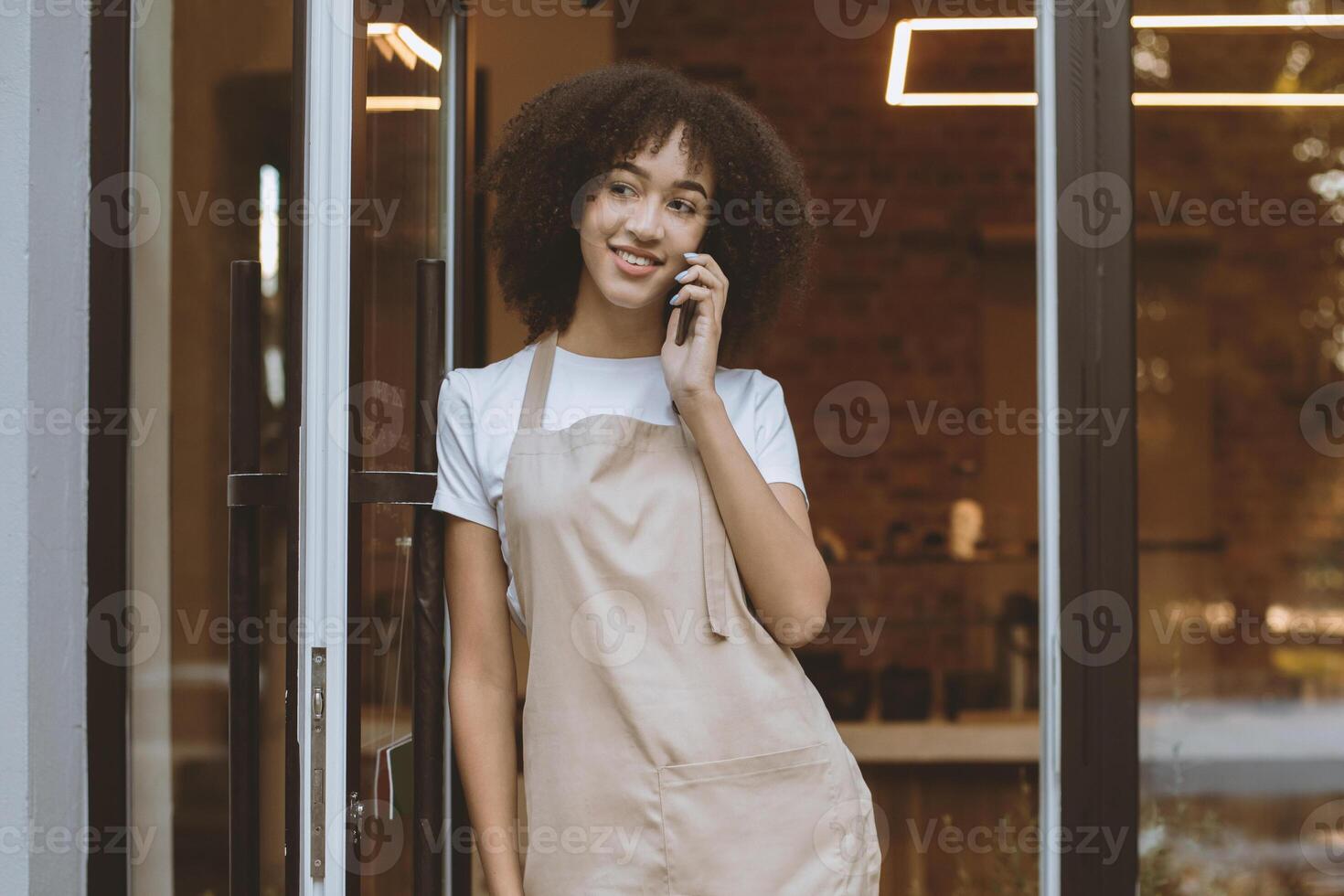 Ordering coffee to go, small business owners and opening after quarantine. Smiling millennial african american woman waitress in apron talking on phone near front door in cafe, free space photo