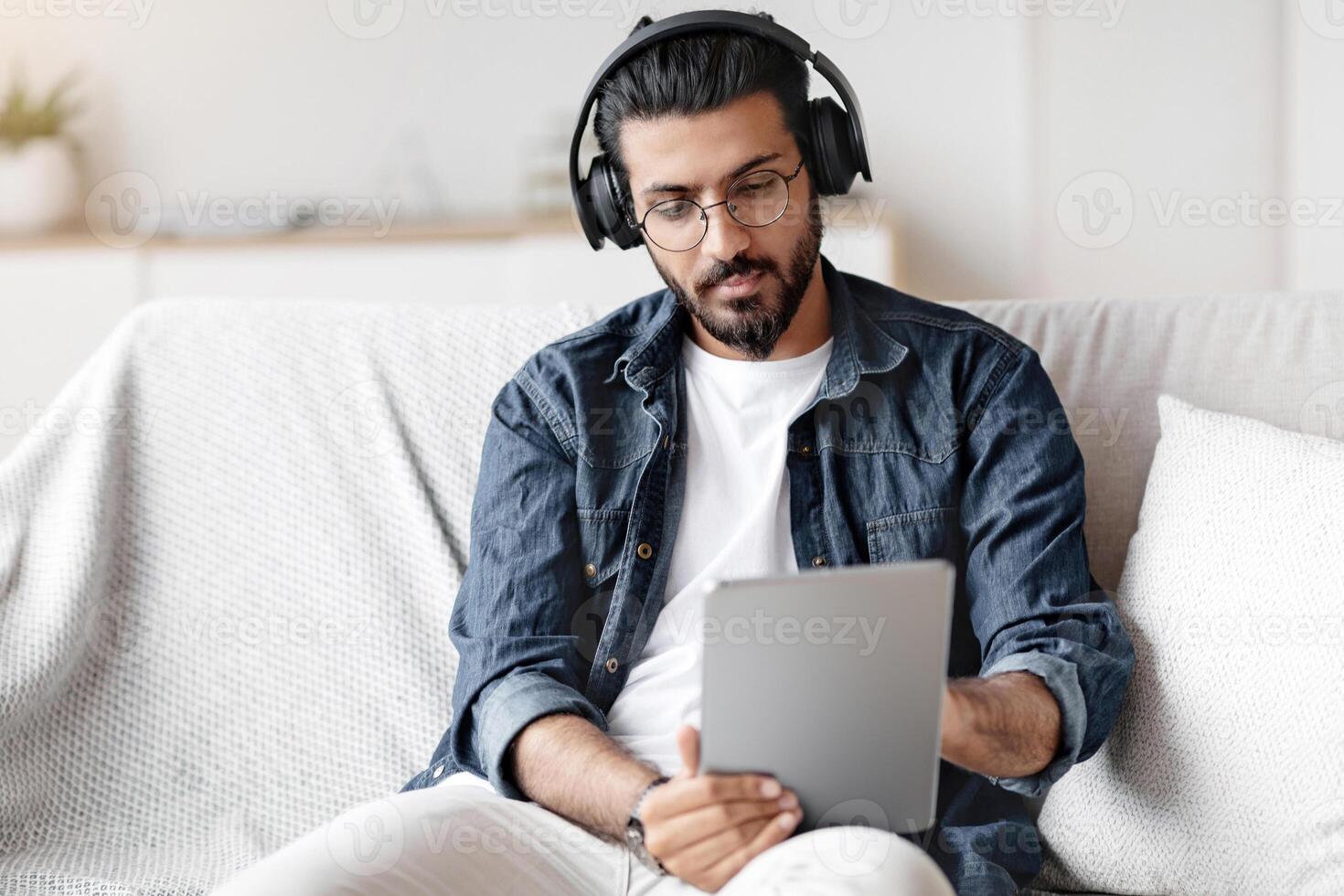 Young arab guy spending time at home with digital tablet and wireless headphones, relaxing on comfortable couch in living room, millenial eastern man checking new application or browsing internet photo