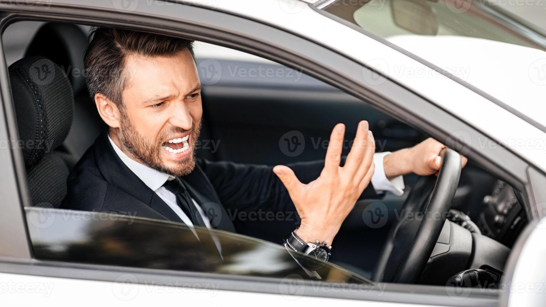 Expressive middle-aged businessman screaming and gesturing while driving car, side view, through window shot. Furious entrepreneur stuck in traffic, getting late for business meeting photo