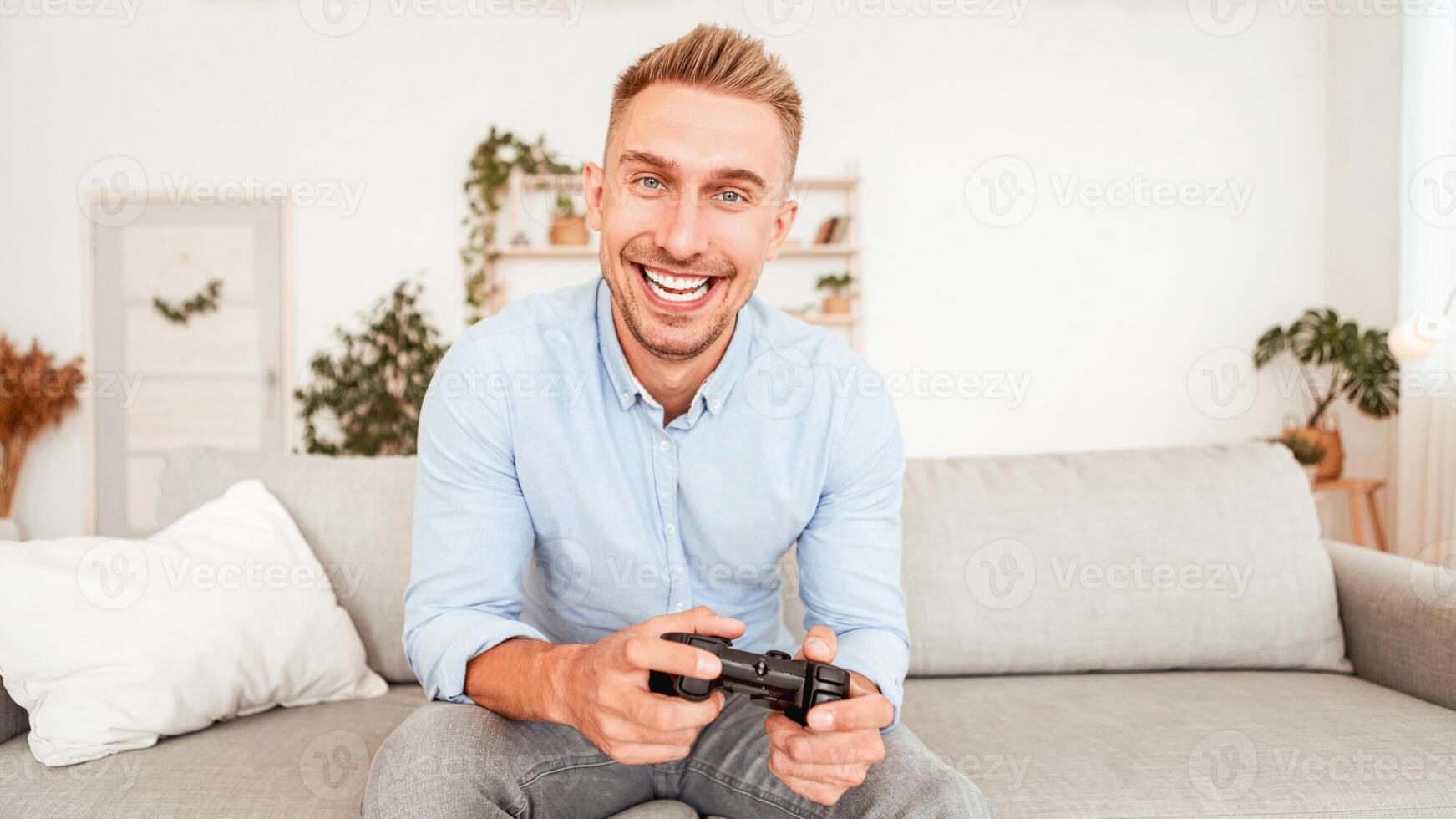 Spending Time At Home Concept. Portrait of happy man playing online games, holding joystick in hands. Adult gamer sitting on sofa at house in living room, looking at camera, blurred background photo