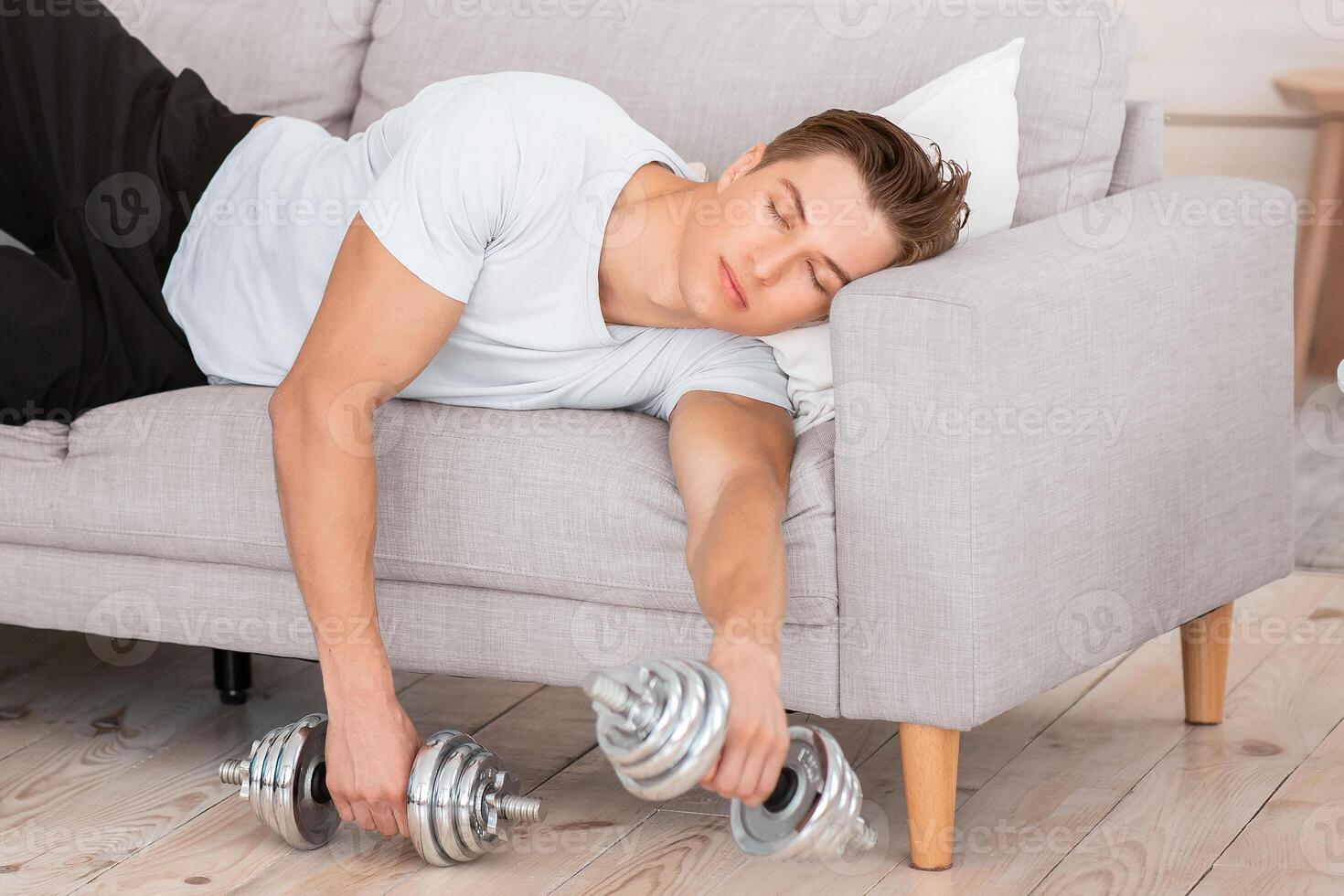 Fatigue after active hard training. Tired guy with dumbbells and fitness ball lies on sofa with closed eyes in living room interior, free space photo