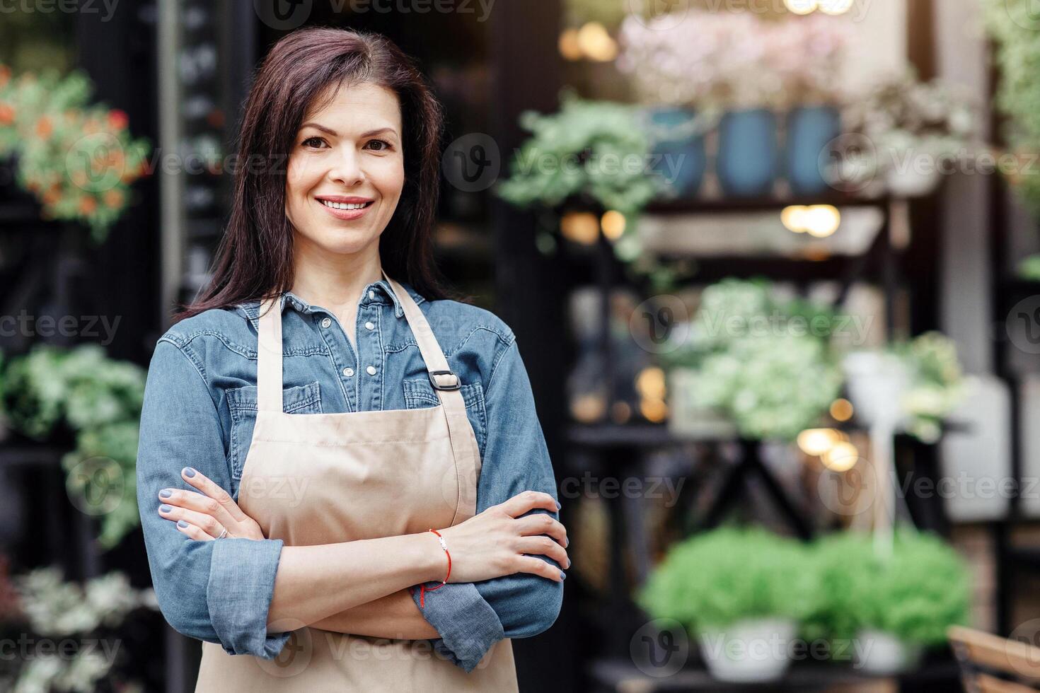 Meeting client and work of seller gardener in flower studio, opening store after covid-19 pandemic. Smiling confident millennial woman in apron with crossed arms near shop with plants, empty space photo