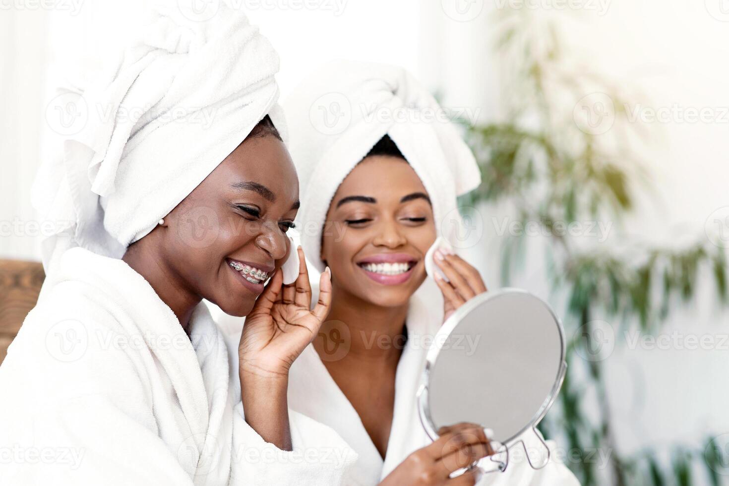 Attractive african american ladies applying toner on faces with cotton pads, looking at mirror. Prertty black women having beauty day at home, looking at mirror and cleansing their faces, copy space photo