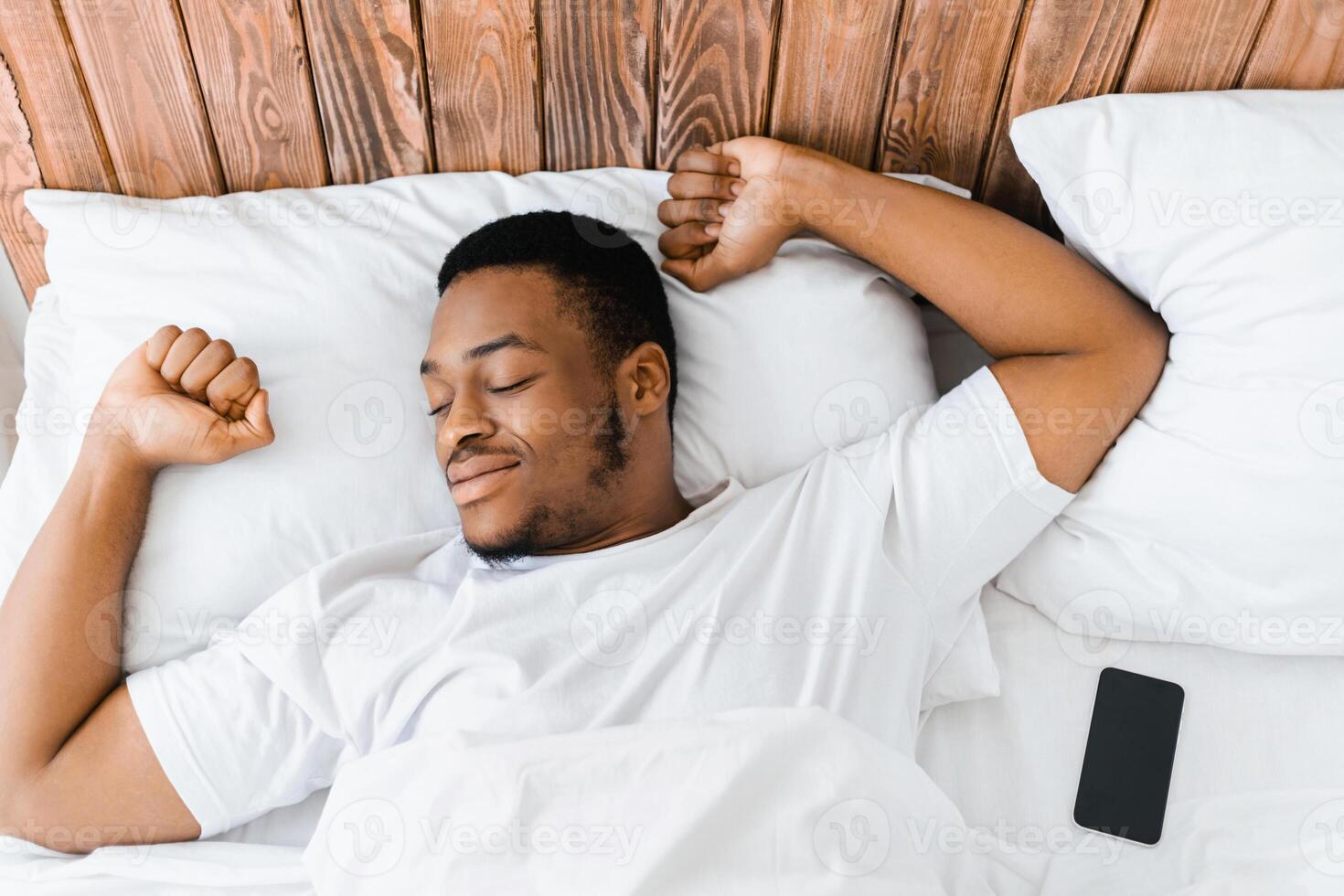 Rested African Guy Waking Up Wellslept Lying In Bed At Home, Stretching Hands Awakening In Cozy Bedroom In The Morning, Smiling With Eyes Closed. Good Rest, Sleep And Recreation Concept. Top View photo
