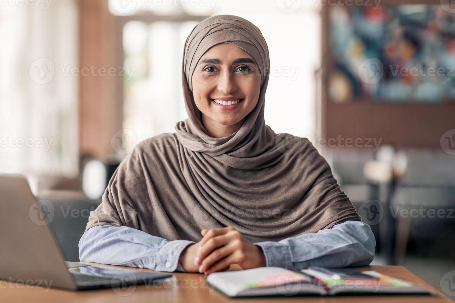 Smiling arab girl looking for job online, sitting at cafe and using laptop, copy space photo