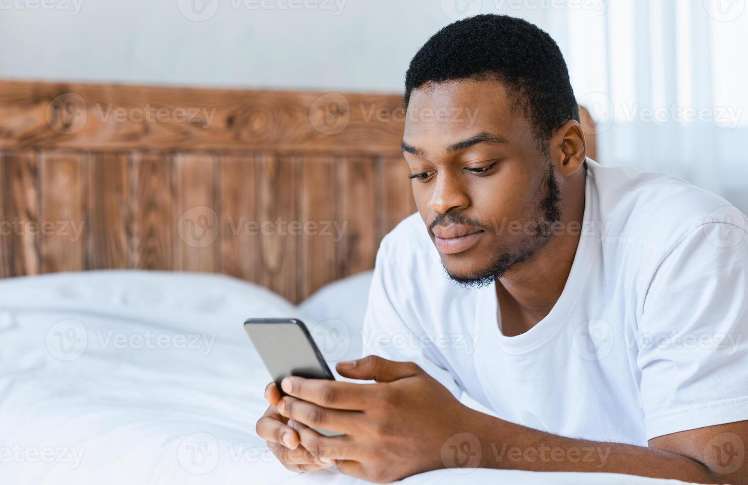 Serious Black Man Using Mobile Phone Browsing Internet Reading Online News On Smartphone Lying In Bed At Home. Cellphone Application Concept. Empty Space For Text photo