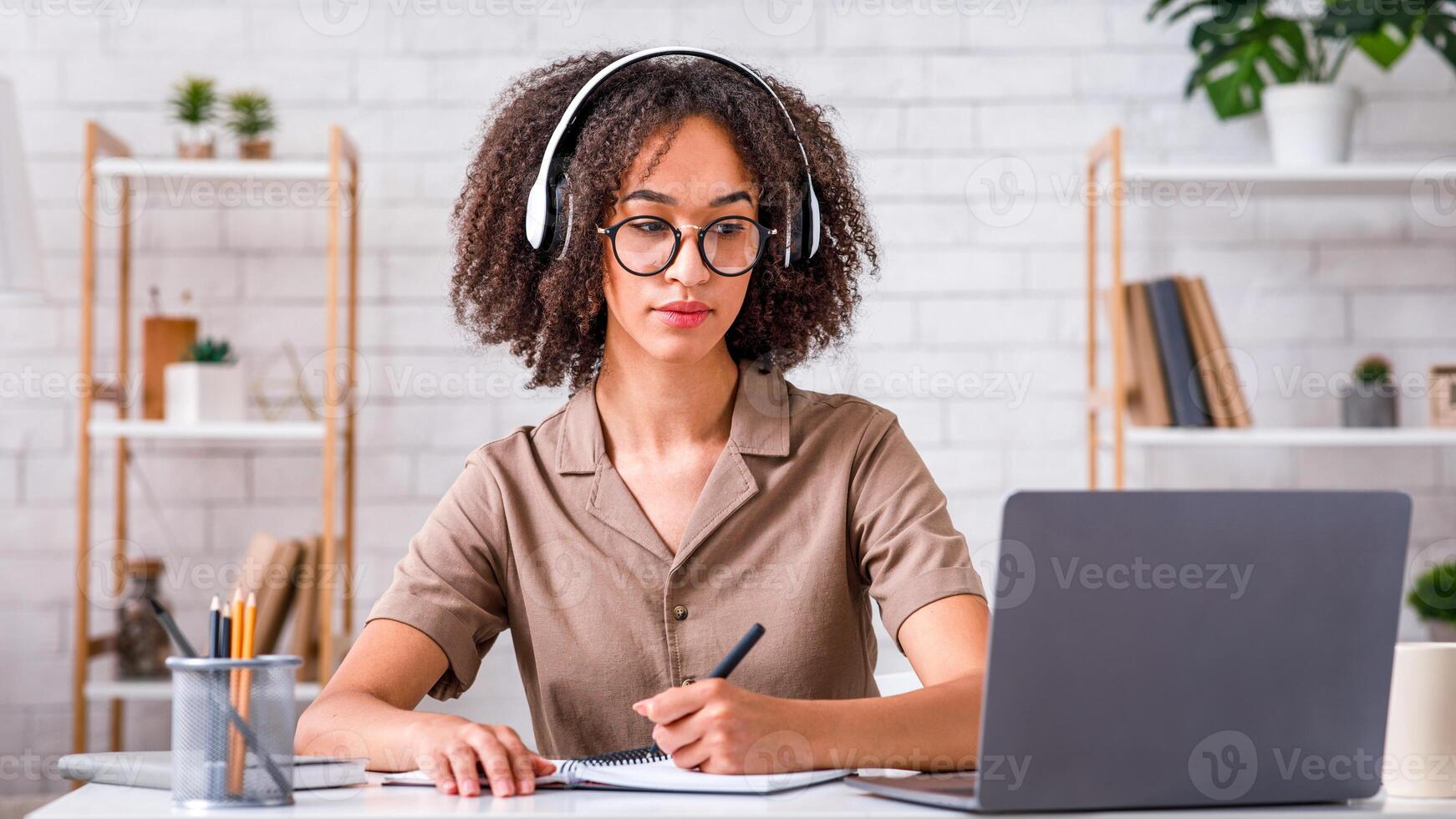 Self-education, online lecture and webinar remotely. Concentrated african american lady in glasses and headphones, making notes in notebook and looks at laptop, at table in living room at home photo