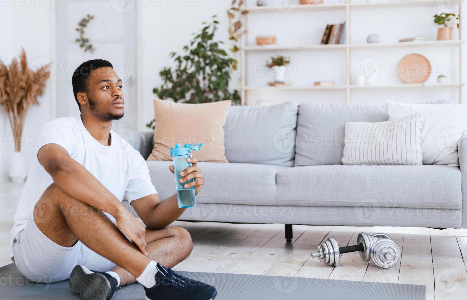 African American Man Holding Bottle Of Water Drinking During Workout Exercising Sitting On Gymnastics Mat At Home. Stay Hydrated, Fitness Hydration Concept. Blank Space For Text photo