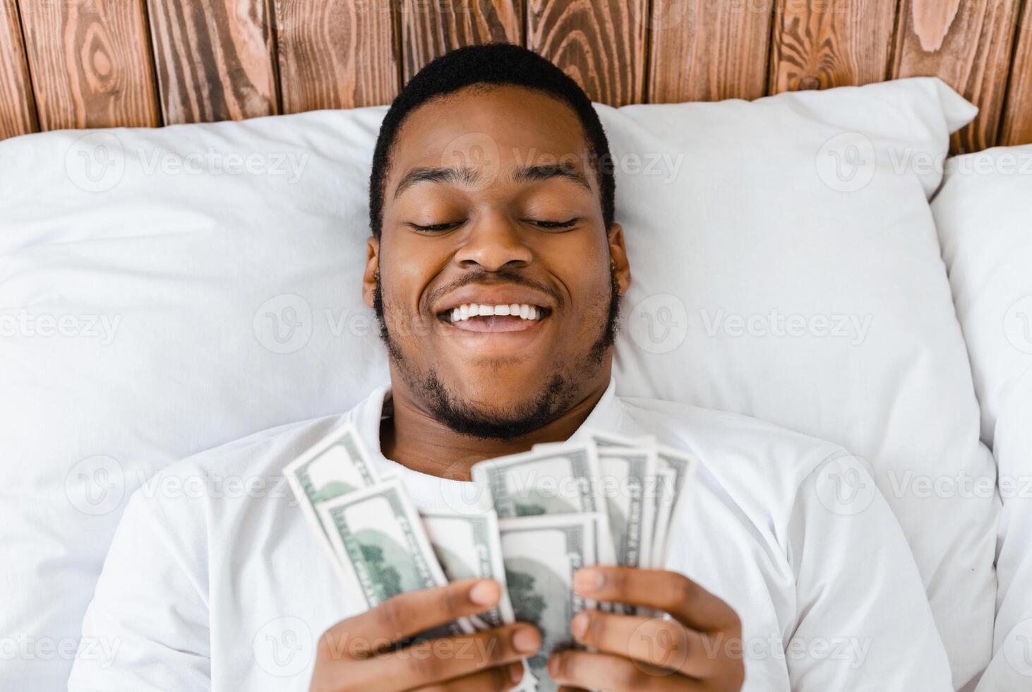 Happy African American Man Holding Money Counting Currency Cash Lying In Bed In Bedroom At Home. Financial Wealth, Budget Income And Finances Savings Safety. Selective Focus, Above View photo