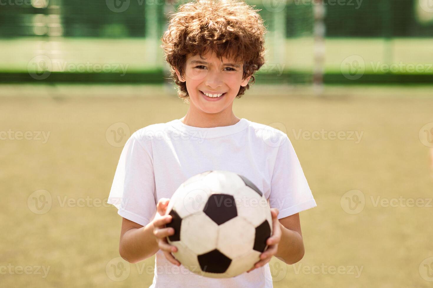 Portrait of cute curly boy holding soccer ball on the football field and looking at camera, posing, selective focus photo