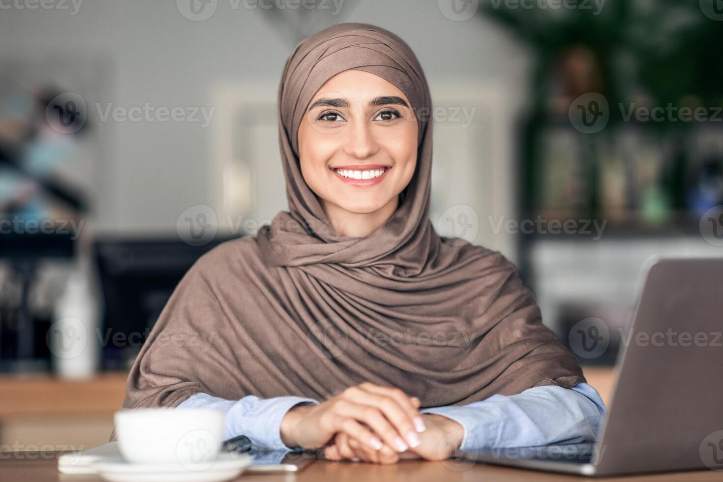 Portrait of positive muslim girl freelancer working online at cafe, using laptop and drinking coffee, free space photo