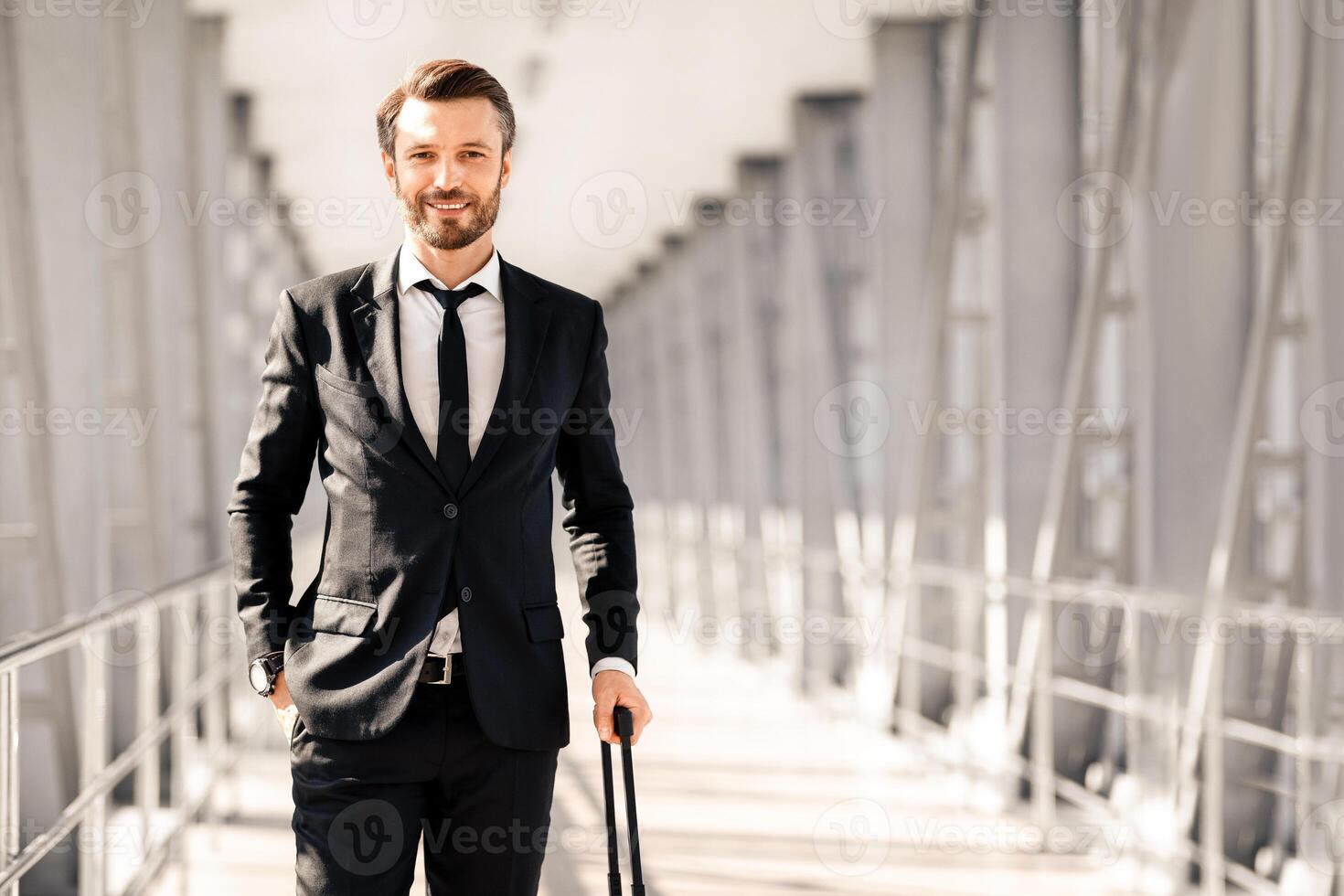 Wealthy middle-aged bearded manager in black suit with suitcase posing in airport, looking at camera and smiling, panorama with copy space. Business trip, business travelling concept photo
