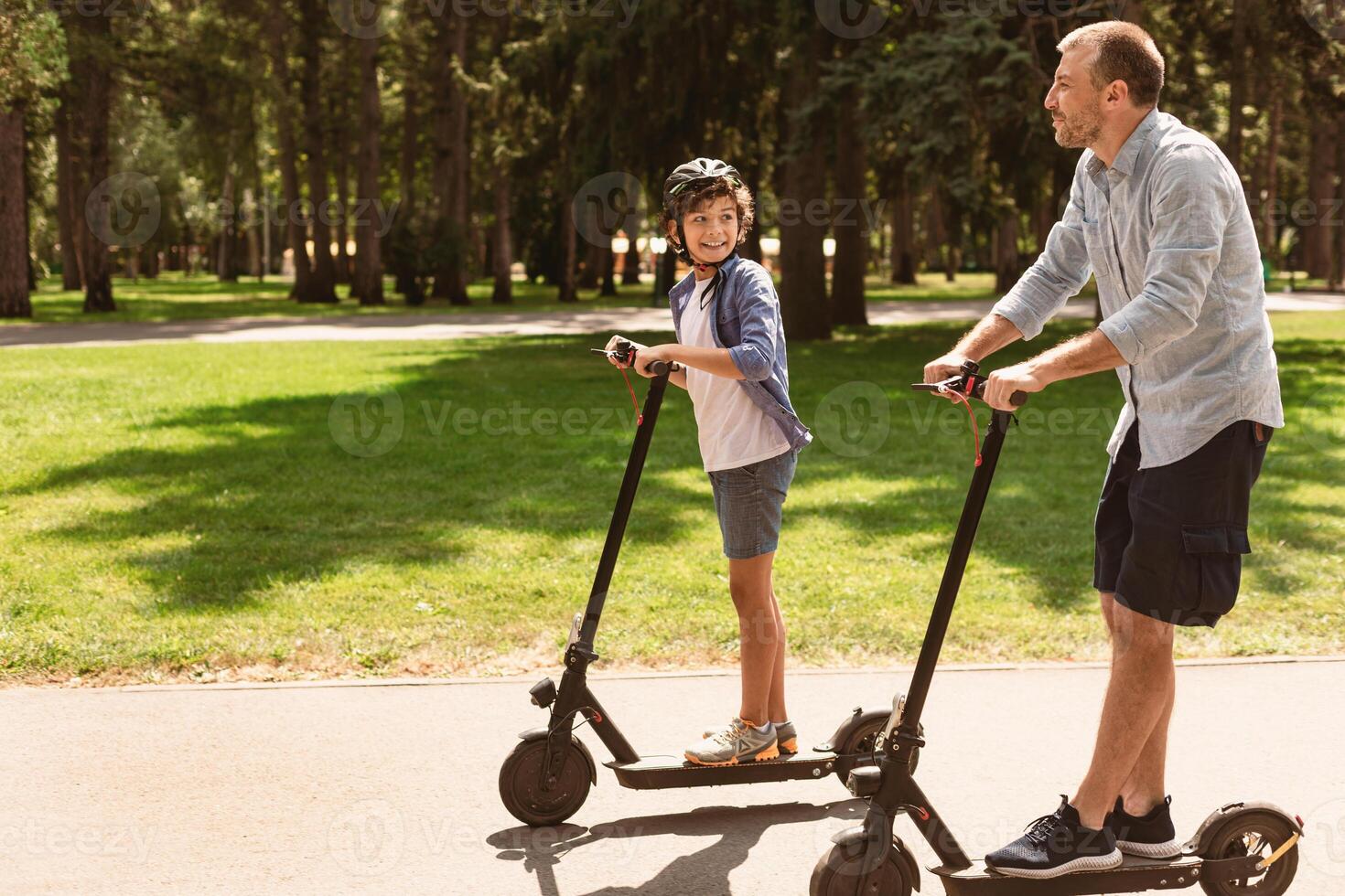 Portrait of happy boy in protective helmet having ride on motorized kick scooter with smiling father at park, spending time together with family on father's day outside, having fun, free copy space photo