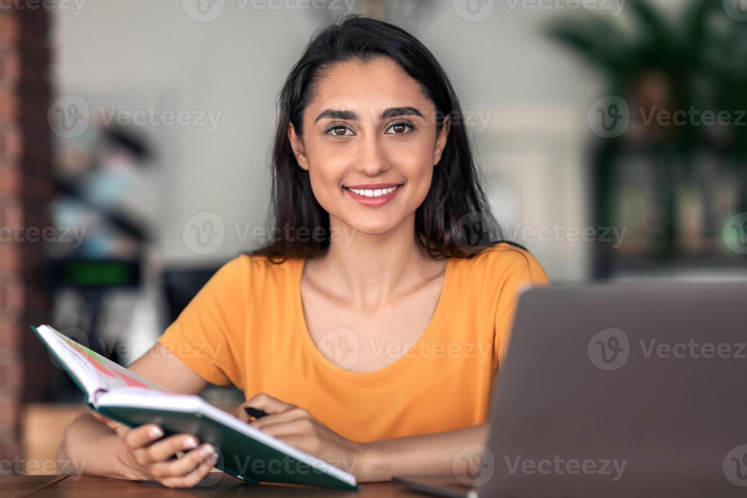 Young cheerful lady looking for part-time job, taking notes at notepad and using laptop at cafeteria, free space. Young arab woman student seeking job opportunities online while resting alone at cafe photo