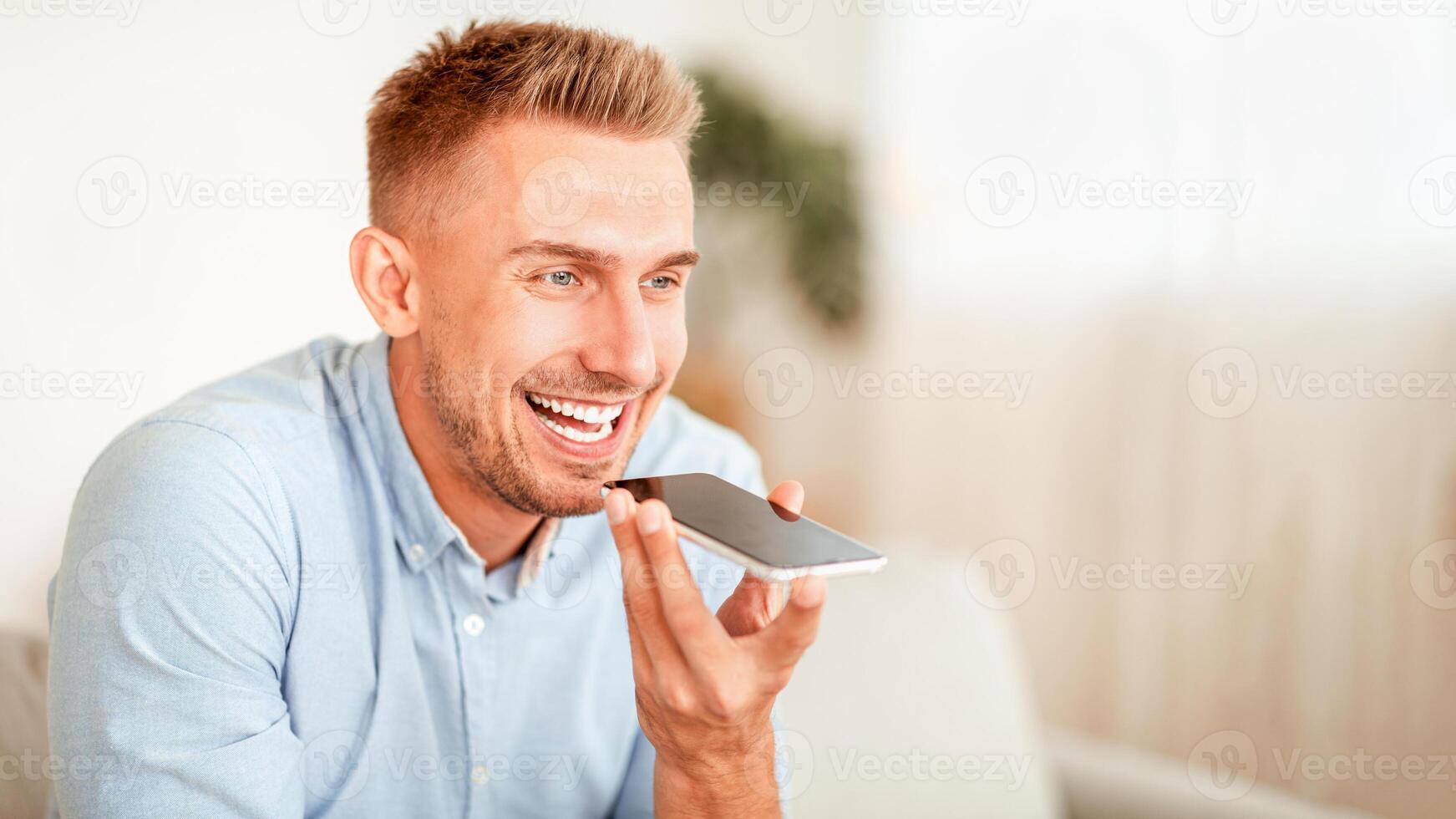 Voice Search Concept. Closeup portrait of young bearded guy using voice assistant or calling on smartphone at home, blurred background, copy space. Smiling man talking on mobile phone photo