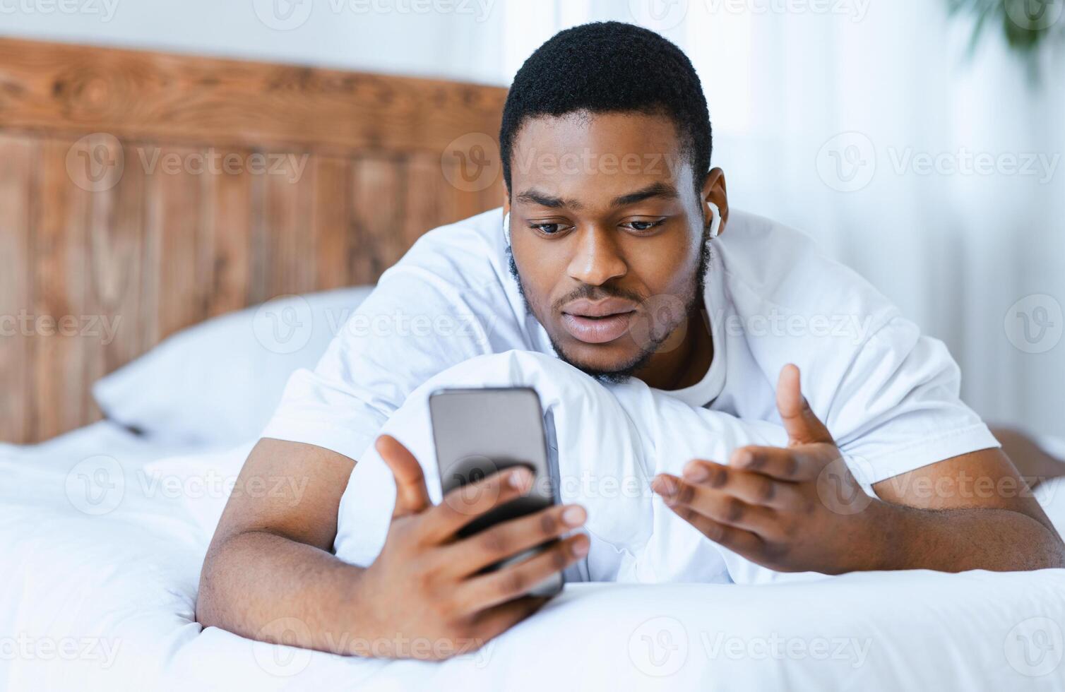 Serious African American Man Making Call On Smartphone Lying In Bed At Home. Distant Online Communication, Conference Mobile Application Concept. Selective Focus photo