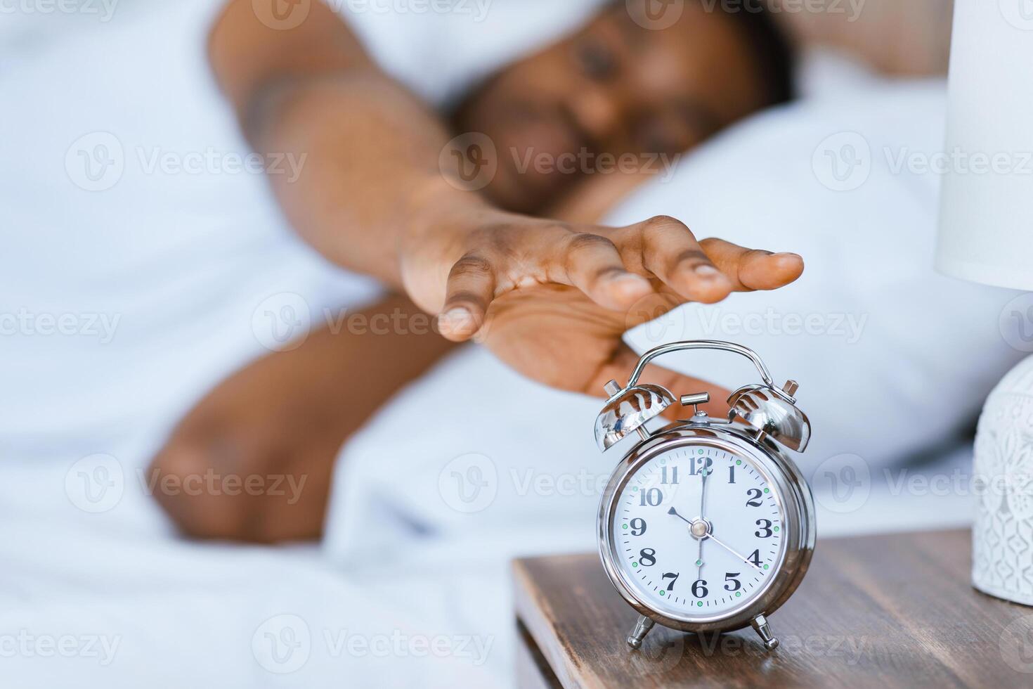 Early Morning Awakening. Sleepy African American Guy Waking Up Turning Off Alarm-Clock Lying In Bed At Home. Time To Get Up Concept. Focus On Alarm Clock, Shallow Depth photo