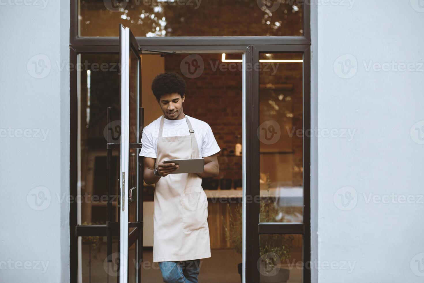 New normal and opening of cafe after covid-19 quarantine. Millennial attractive african american man in apron working on digital tablet near front door of modern cafeteria, empty space photo