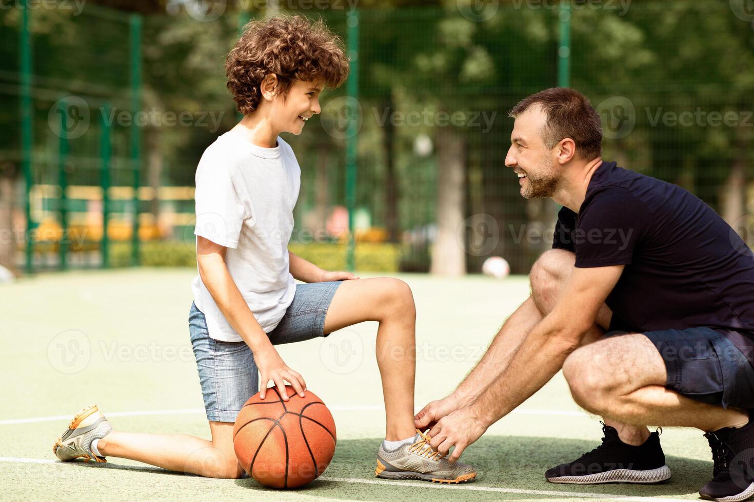 Preparation. Portrait of supportive man tying shoelaces of curly boy on basketball playground, blurred background photo