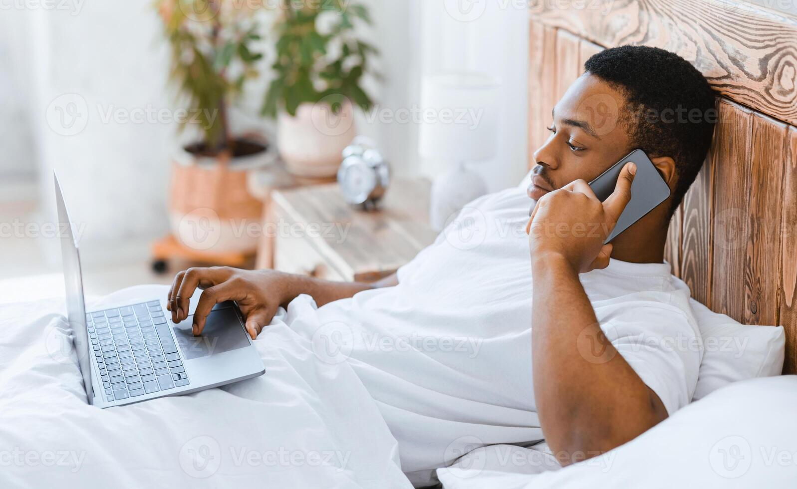 African American Man Talking On Cellphone Receiving Phone Call Working On Laptop Computer Lying In Bed At Home. Freelance Lifestyle, Always In Touch, Mobile Communication Concept. Selective Focus photo