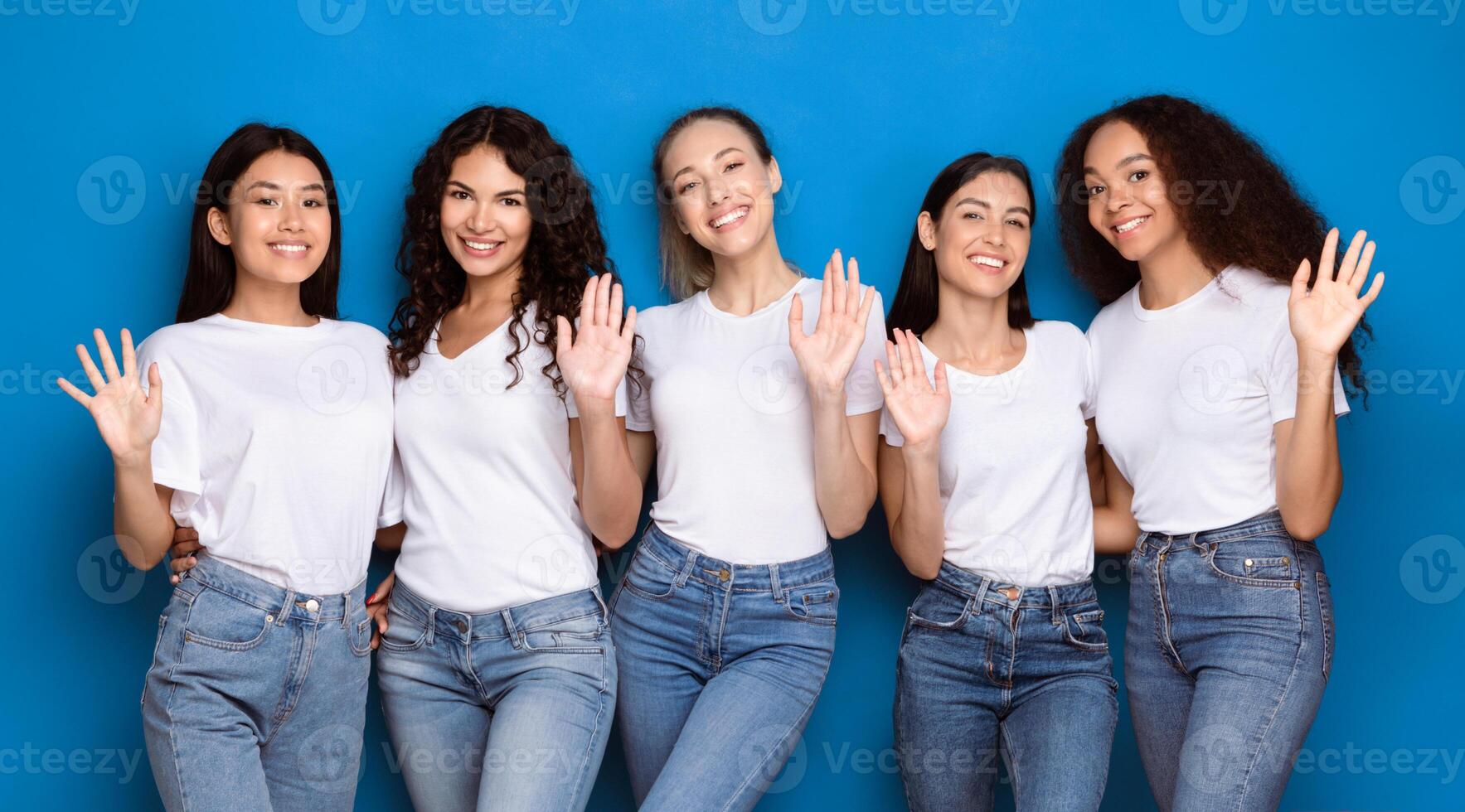 Hello Greeting. Five Multiethnic Women Waving Hand Smiling To Camera Posing On Blue Background. Cheerful Diverse Ladies Gesturing Hi Standing In Studio, Wearing Casual Clothes. Panorama photo
