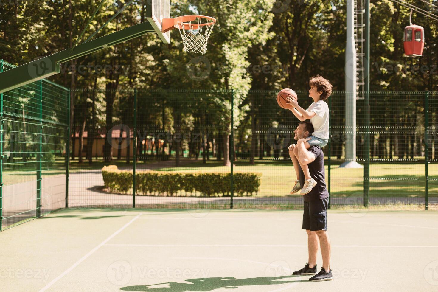 Sports Concept. Curly boy sitting on the man's shoulders, throwing ball into basket, side view photo