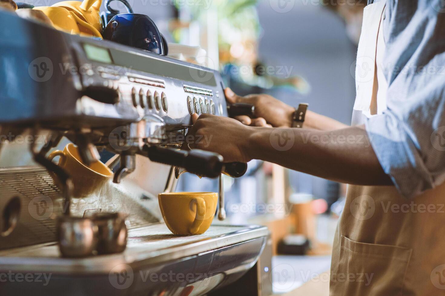 New reality, modern work during covid-19 lockdown, small business and startup. Young african american male in apron makes latte in yellow cup on professional coffee machine in cafe interior, cropped photo