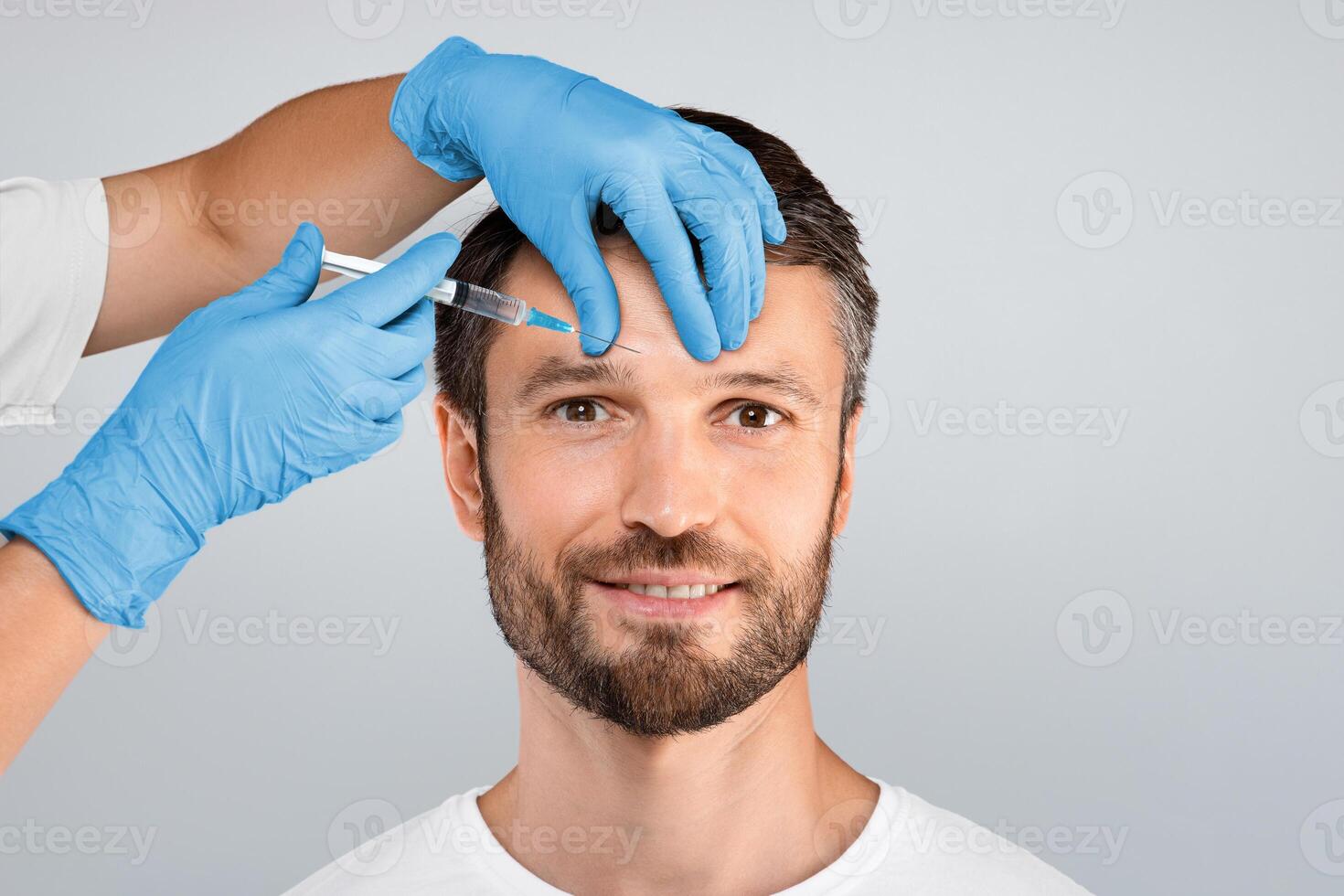 Attractive middle aged bearded man getting brow lift over grey studio background, copy space. Unrecognizable beautician or plastic surgeon making beauty injection for handsome smiling man photo