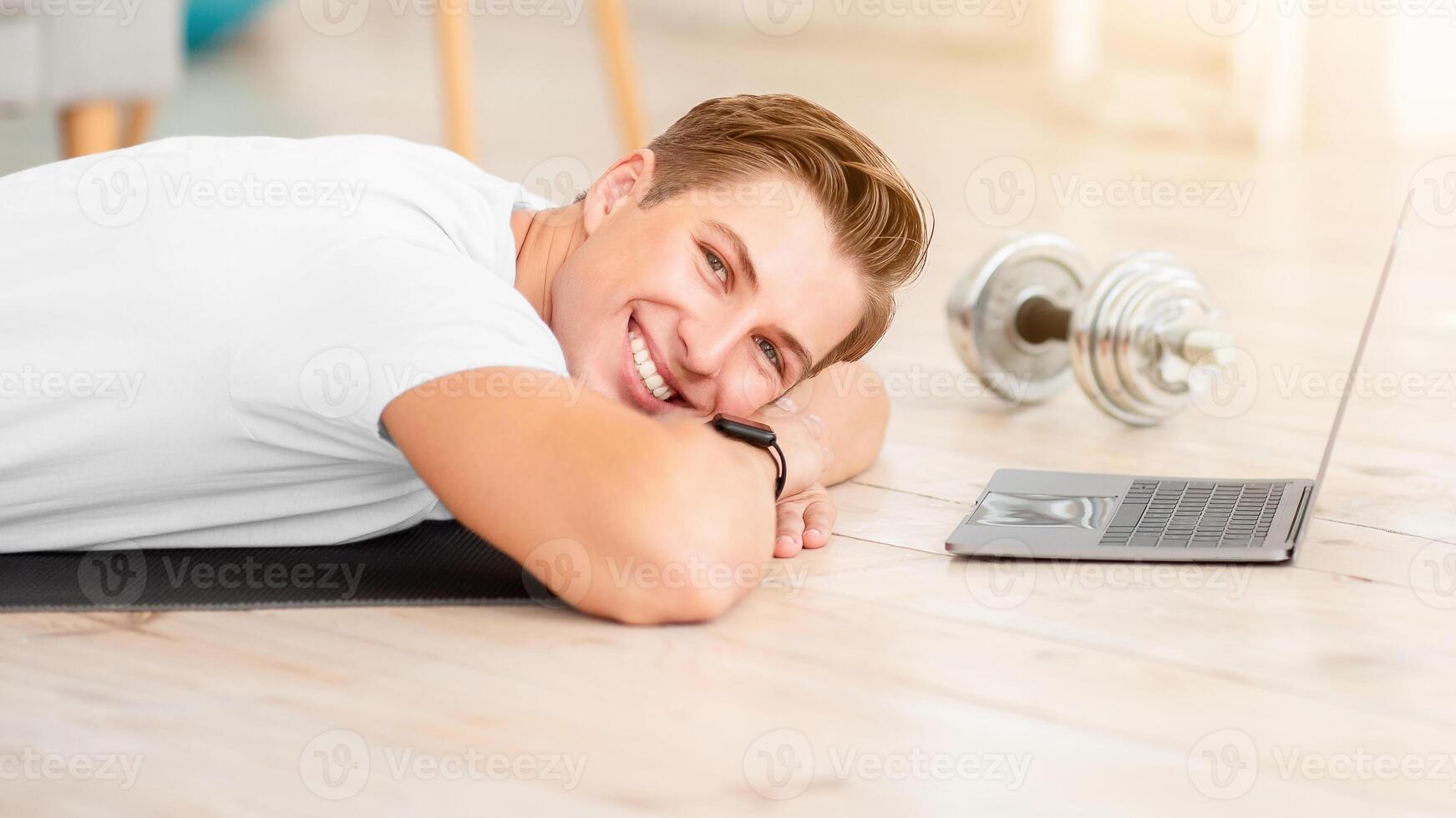 Online workout and rest. Smiling man lying on floor on mat in interior of living room with laptop, cup for protein shakes and dumbbells, panorama, free space photo