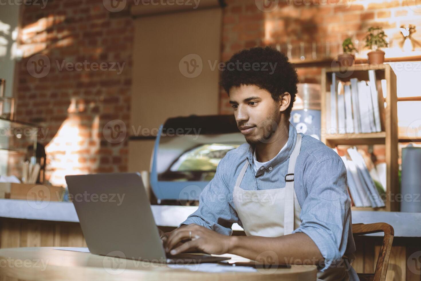 Man owner working and checking email on computer. Serious young african american guy in apron use laptop and social networks in modern loft cafe in morning after lockdown, free space photo