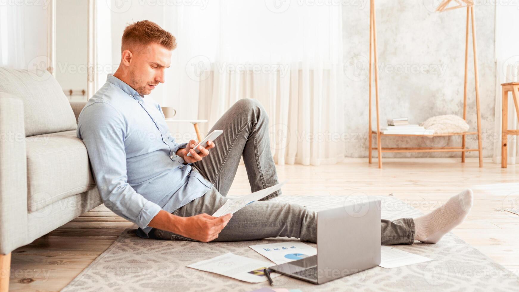 Young freelancer working from home, sitting on floor with pc, using smart phone and holding documents photo