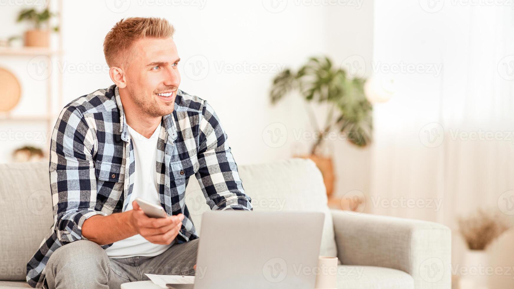 Home Office. Happy casual man holding smartphone and using laptop, daydreaming and looking away at copy space. Successful professional guy working remotely and thinking about business and future photo